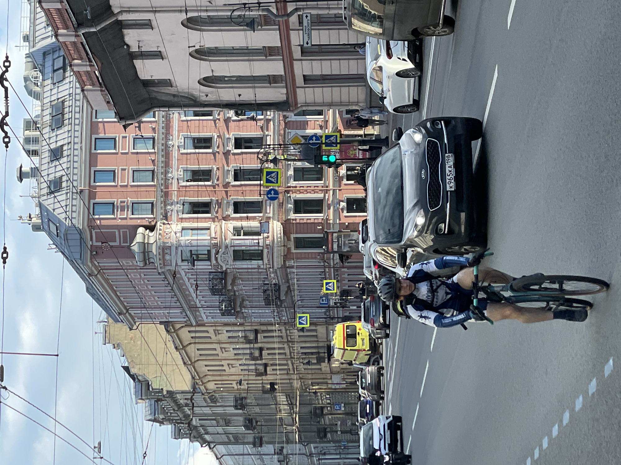 The image captures a street scene in Saint Petersburg, Russia, presenting a cyclist riding in the traffic flow alongside cars on a wide, multi-lane road lined with historic buildings. The architecture is characteristic of the city, with elaborate facades, decorative windows, and ornate detailing. Traffic signals and pedestrian crossing signs are visible, suggesting a bustling urban environment. The presence of a yellow ambulance suggests an emergency service vehicle moving through the traffic. In the foreground, a cyclist wearing a helmet and riding gear is navigating the road. Cars of different colors and models surround the cyclist. The license plate of the black Kia reads "K965KA 198". Overhead wires crisscross the sky, common in urban settings for public transportation.