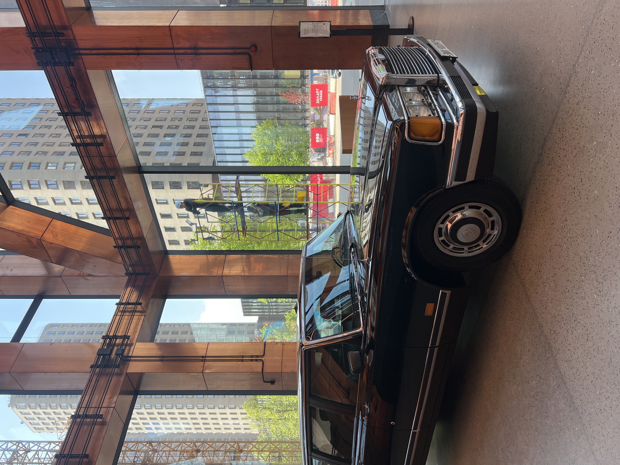 The image captures a dark, classic ZIL limousine parked indoors, likely in a museum or exhibition space in Moscow, Russia, given the "ЗИЛАРТ ГРАД" banner visible through the large windows. The car is positioned close to the window, reflecting the surrounding cityscape, including modern buildings and construction activity. The interior architecture features exposed wooden beams and metal supports, giving an industrial-chic aesthetic. No people are directly visible, but a distorted reflection in the car's window suggests human presence in the outside environment. The scene is well-lit, highlighting the car's shiny exterior and the contrast between the indoor display and the bustling city outside.