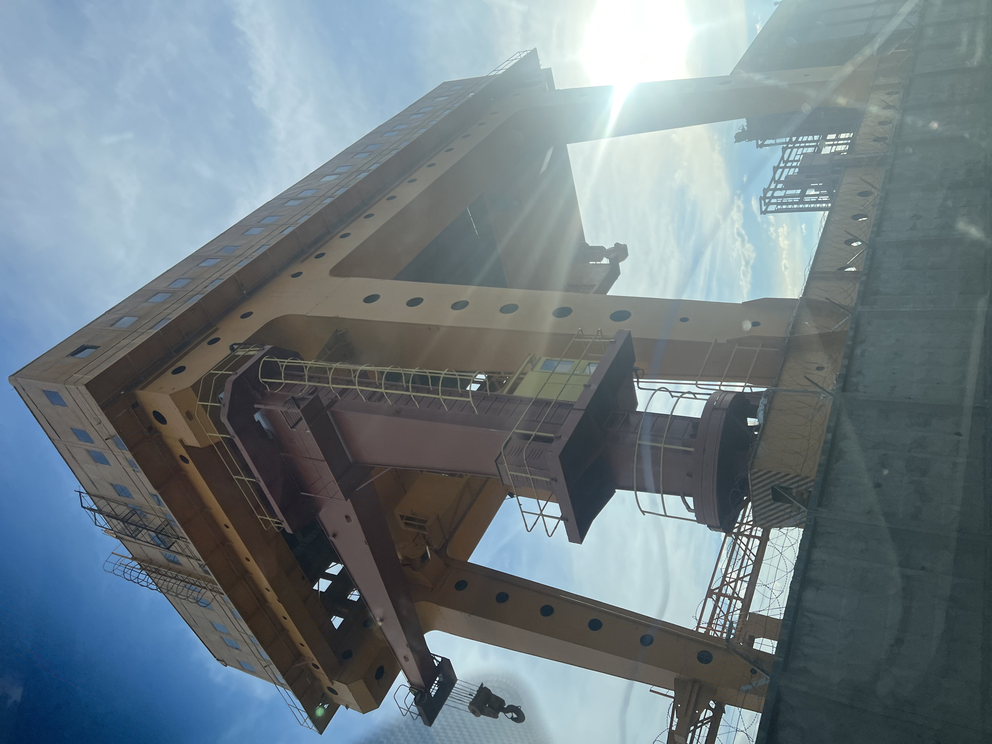 The image depicts a large gantry crane in the city of Chaikovsky, Russia. The crane is predominantly yellow and brown, with a tall, rectangular structure featuring evenly spaced windows and circular openings. A central vertical component with a cab is suspended within the crane's frame. Metal support beams with attached ladders and safety railings are visible. A hook and cable dangle from the lower section. The sky is blue with light clouds and the sun is positioned above the crane, creating lens flare in the image. The bottom right of the frame shows a concrete barrier topped with barbed wire. The image is taken from a low angle, looking upwards at the crane.