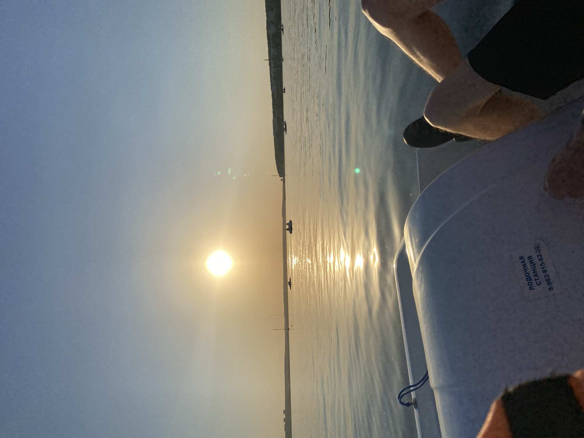 The image depicts a sunset scene on a body of water, presumably a lake or river, with the sun low on the horizon. A coastline is visible in the distance. The viewer is likely positioned on a boat, as the front of a white boat and legs of a person are visible in the frame. The boat has an inscription in Russian, which appears to read "Лодочная станция 8-982-815-82-32," meaning "Boat Station" followed by a phone number. Based on the location of the boat station and the phone number prefix, the image is likely from Izhevsk, Russia.
