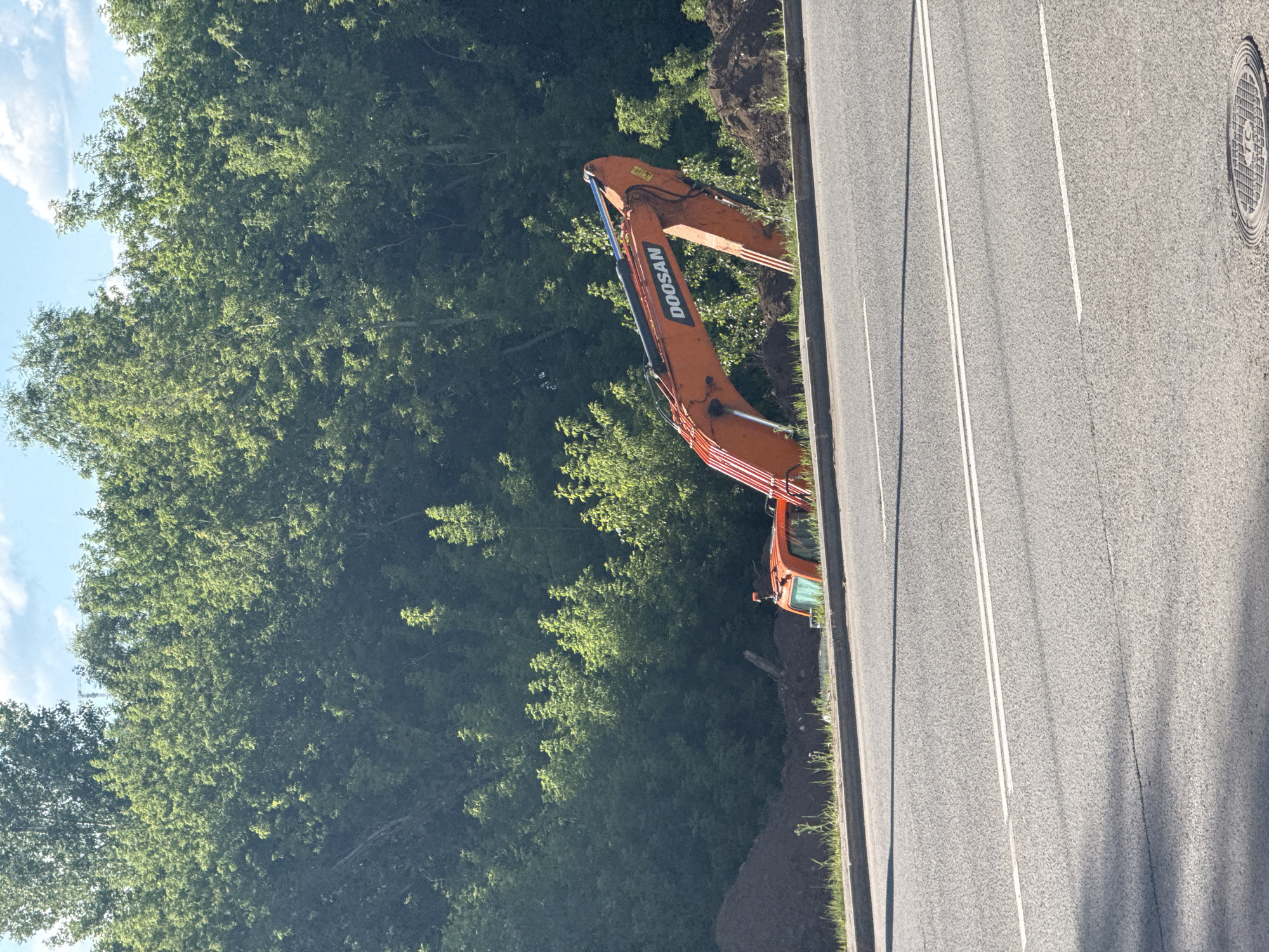 The image shows an orange excavator next to a road. The excavator has the brand name "DOOSAN" printed on its arm. The road has white lane markings. To the left of the road is a dense green forest. The sky is blue with some white clouds. The sun appears to be shining, casting shadows. The scene seems to be a construction or road work site. There are no people visible in the image.