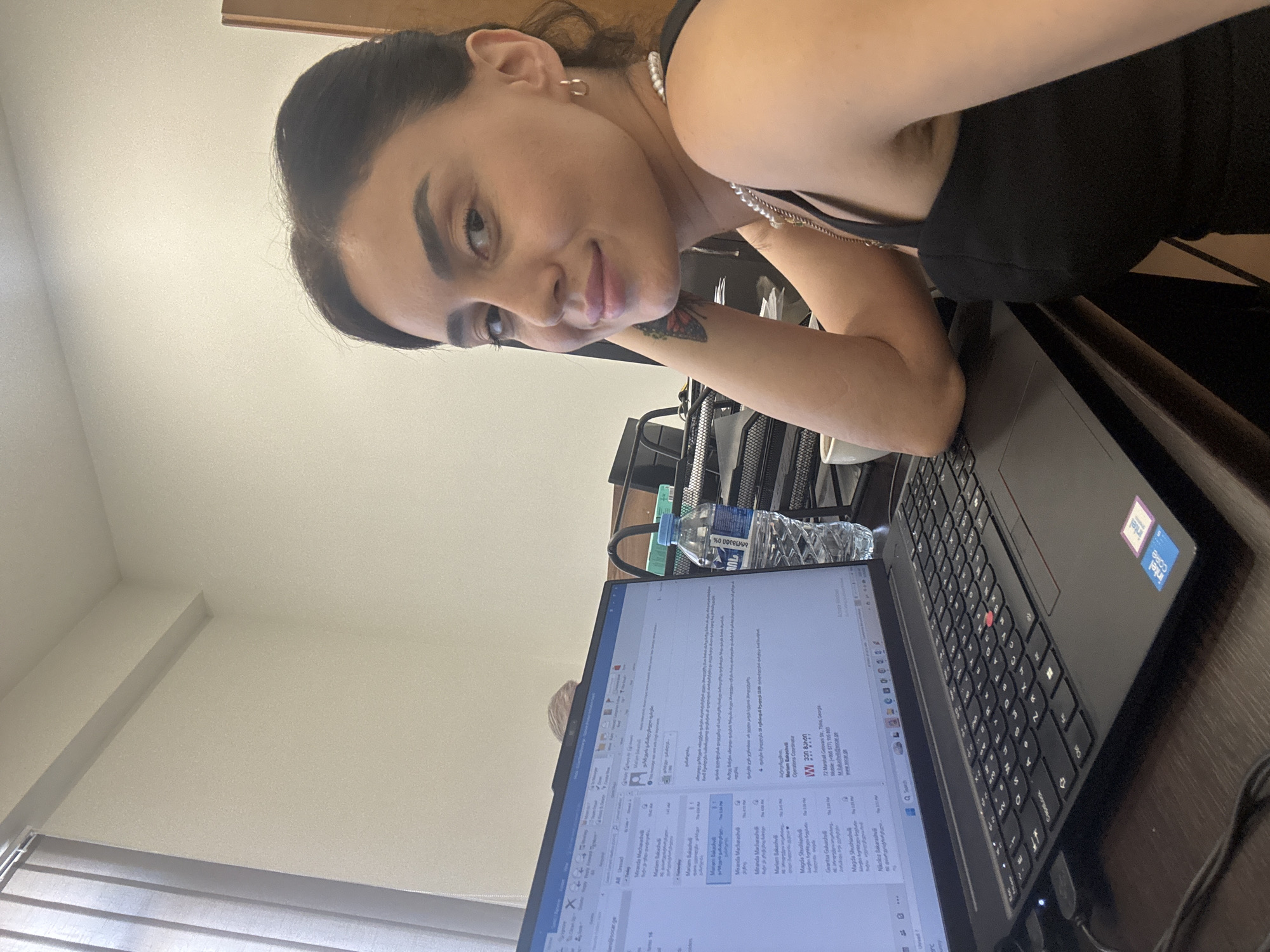 A woman with dark hair and dark eyes is sitting at a desk in an office in Tbilisi, Georgia. She is looking at the camera with a slight smile. She is wearing a black top and a pearl necklace. A laptop is open in front of her, displaying an email interface. A bottle of water is on the desk next to the laptop. The room is lit by natural light coming from a window with vertical blinds on the left side of the image. The desk is made of dark wood.