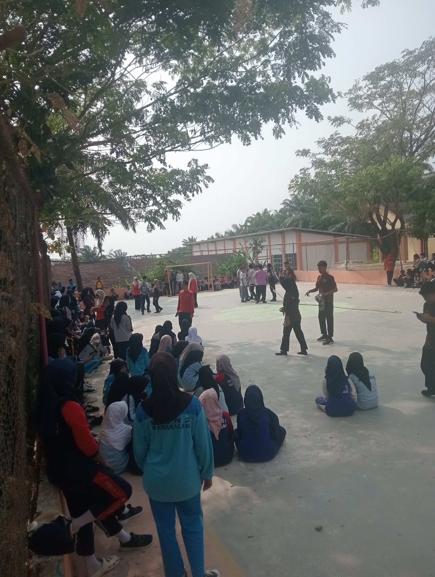 The image depicts an outdoor scene in Wanasalam, Indonesia, possibly at a school or community gathering. A group of people, predominantly young individuals, are gathered on a paved area. Many are seated on the ground, while others are standing. Some individuals are wearing headscarves. The environment includes trees with dense foliage, a building in the background with orange trim, and a chain-link fence on the left. The ground appears to be a light-colored concrete or asphalt surface. There is a noticeable presence of sunlight, suggesting it is daytime. Some individuals appear to be observing an activity, while others are engaged in conversation or rest. The overall atmosphere suggests a casual social event or a break period.