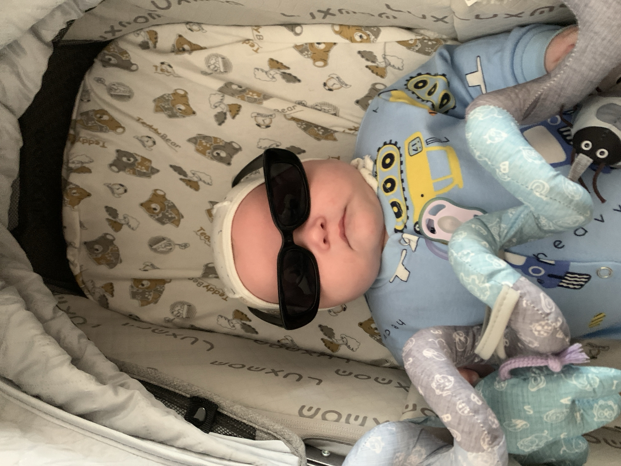 An infant wearing a white beanie and black sunglasses is lying in a stroller. The stroller is lined with a fabric featuring a pattern of teddy bears. The infant is dressed in a blue onesie with a yellow construction vehicle design. There are plush toys in the stroller with the infant. The scene is indoors.