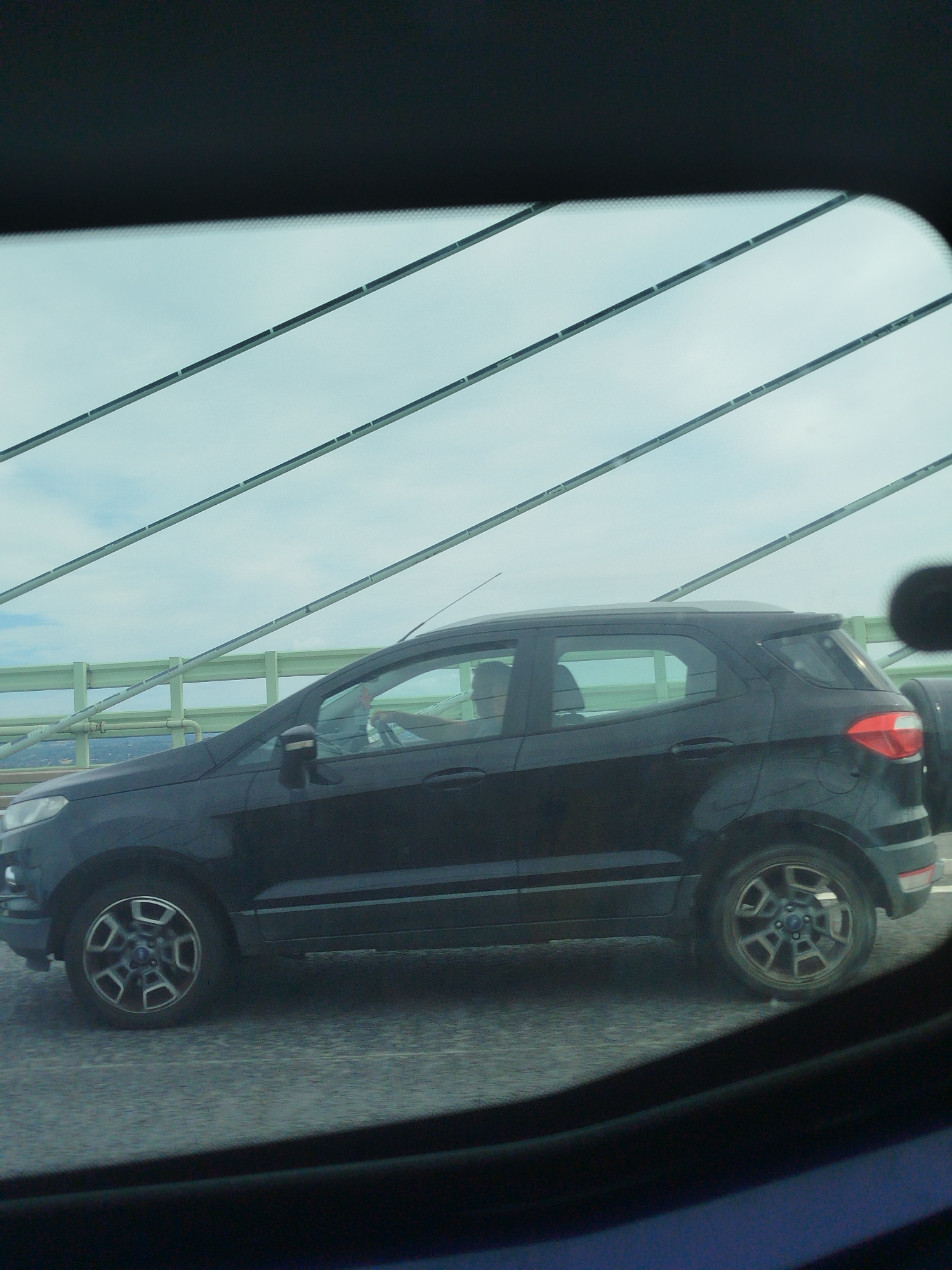 A black Ford EcoSport SUV is captured in profile, driving on a bridge. The driver, a person with short dark hair and a light-colored shirt, is visible behind the steering wheel. The vehicle is moving in the foreground, with the driver's side facing the viewer. The bridge structure is prominent, with diagonal cables and a green guardrail visible in the background. Beyond the bridge, a hazy cityscape with buildings can be observed. The sky above is overcast with a pale blue hue. The image appears to have been taken from inside another vehicle, as suggested by the darkened frame and reflections in the foreground. The specific location is on the Caldicot Pill Viaduct, which is part of the M48 motorway in Caldicot, United Kingdom.