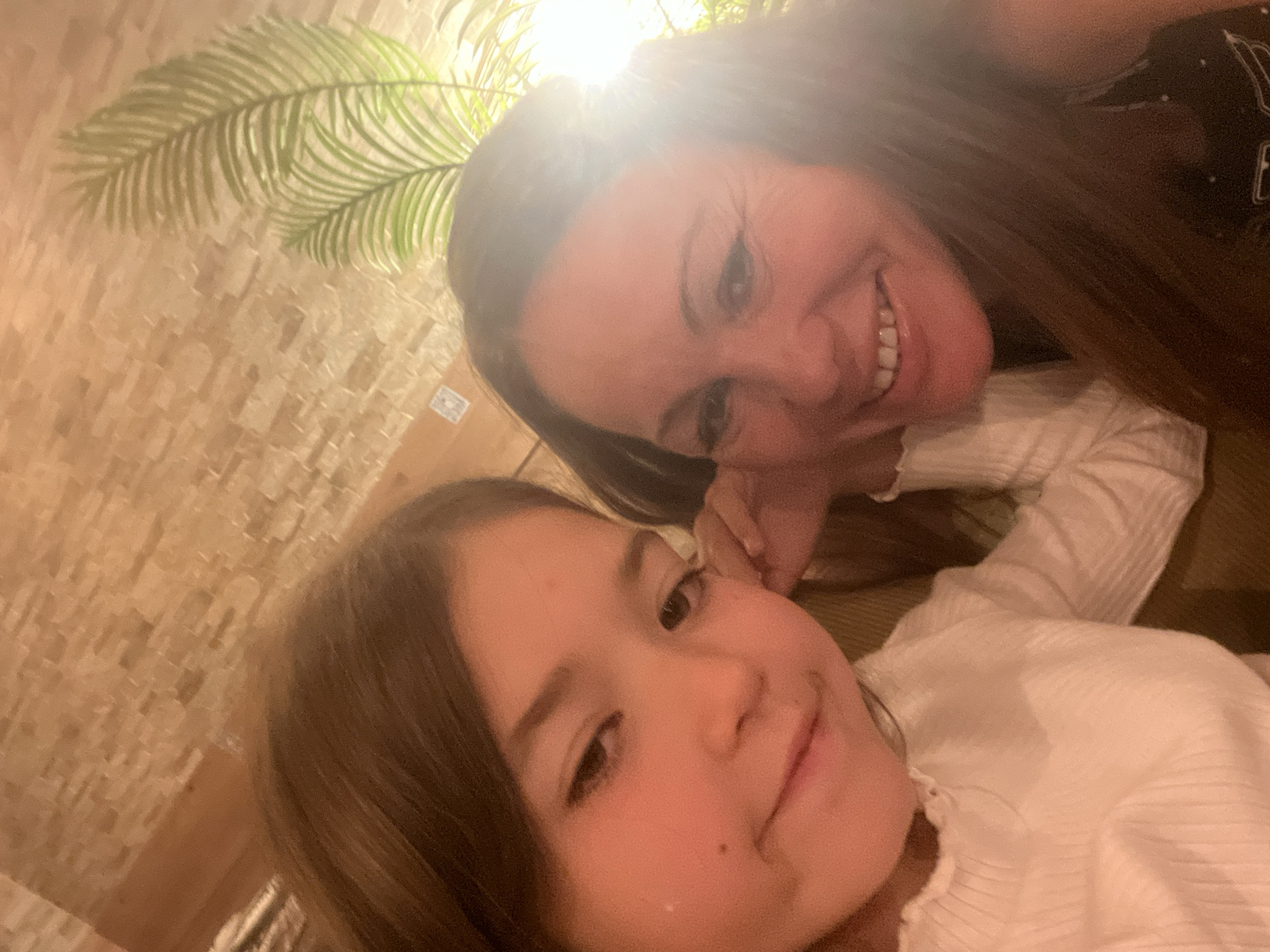 A woman and a young girl are in a close-up selfie, possibly indoors, with a decorative palm frond visible in the upper left corner. Both are smiling. The woman has long dark hair and is wearing a dark top. The girl has brown hair and is wearing a white top. The background appears to be a wall with a textured or patterned surface. The location is Sant Pere I Sant Pau, Spain.