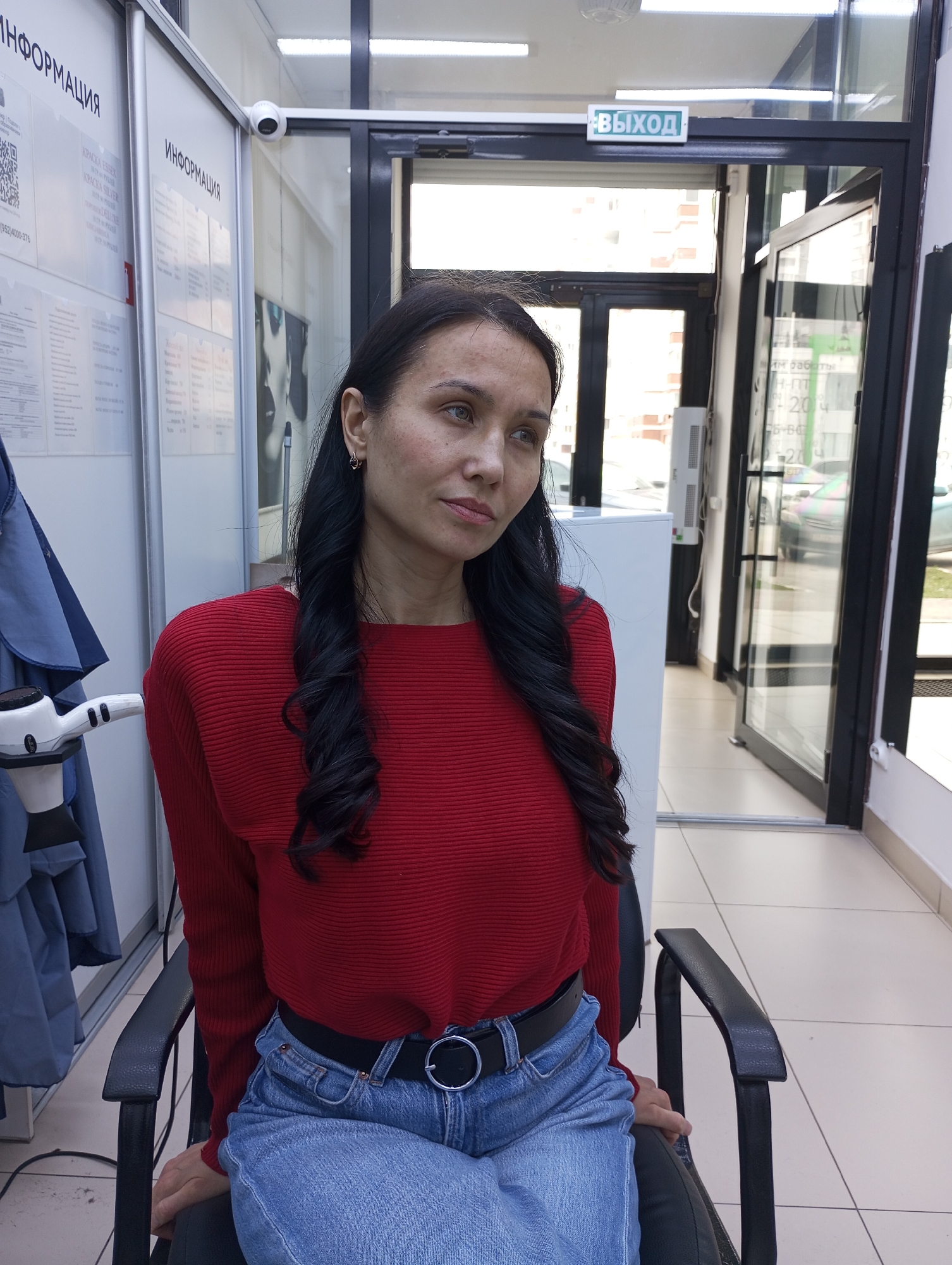 An indoor, eye-level portrait shows a woman seated in a dark chair, looking slightly to her right. She has long, dark, wavy hair and is wearing a red long-sleeved top, light blue jeans, and a black belt with a silver buckle. Her expression is neutral to pensive.
The setting appears to be a commercial space, likely a salon or waiting area, with light-colored walls and floor tiles. To the left, a blue smock or cape hangs near a white stand holding a professional hair dryer. In the background, white informational boards are mounted on the wall. A security camera is visible near the top left corner.
To the right, a large glass door serves as an exit, allowing natural daylight to illuminate the scene. Cars and buildings are faintly visible through the glass, indicating an outdoor street view during the daytime.
Visible text on the informational boards includes "ИНФОРМАЦИЯ" (Information) repeated twice, "КРАСКА ESSEX", "KPACKA SILVER", and "DELUXE" (likely product names), and a phone number "(952)4000-376". Above the exit door, a green sign clearly reads "ВЫХОД" (Exit). Another sign near the door shows "им работы" (work hours) along with numbers like "2004" and "09-95".