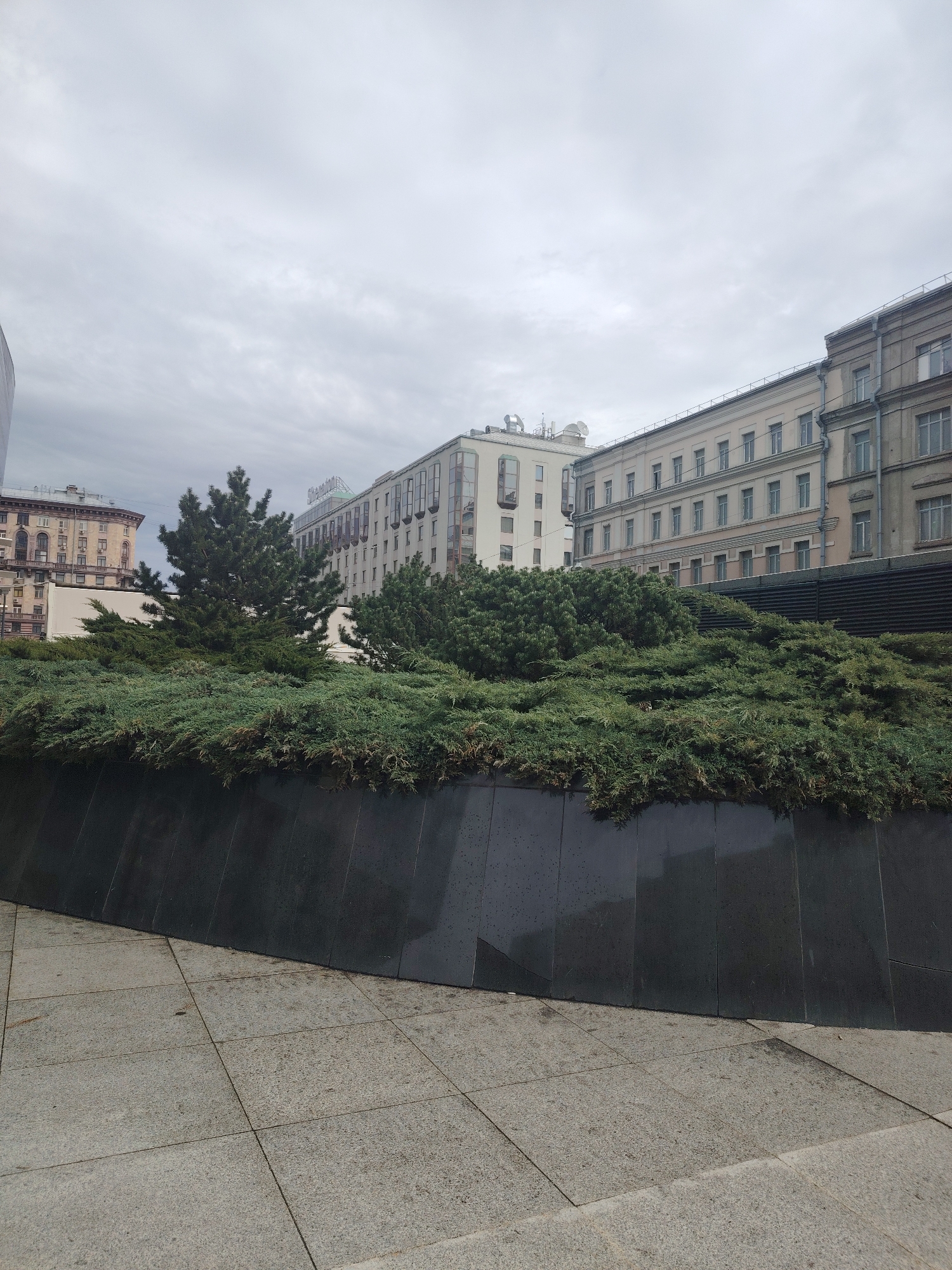 This outdoor urban scene, captured on an overcast day under a uniform grey sky, showcases a blend of historic and modern architecture in Moscow, Russia.
In the foreground, grey square concrete pavers cover the ground, leading to a dark, possibly granite, curved wall that forms a raised planter. This planter is densely filled with lush green, low-lying evergreen shrubs, likely juniper, and a few taller conifer trees with dark green needles.
Beyond the greenery, several buildings define the midground and background. To the far left, a portion of an older, multi-story building with ornate facades and arched windows is visible. Dominating the center-right is a long, light-colored building with rows of windows and balconies. On its roof, a prominent white sign with green lettering clearly displays "BALTSCHUG KEMPINSKI," identifying the well-known Baltschug Kempinski Hotel. Further to the right, another multi-story building with a lighter facade and classic architectural lines stands adjacent to a slightly darker, older building, both featuring numerous windows.
No people are visible in the image, and no specific activities are taking place. The time of day appears to be daytime, though the heavy cloud cover suggests a dull or possibly chilly atmosphere. The overall impression is one of a quiet city street or plaza, emphasizing the architectural heritage and the green landscaping.