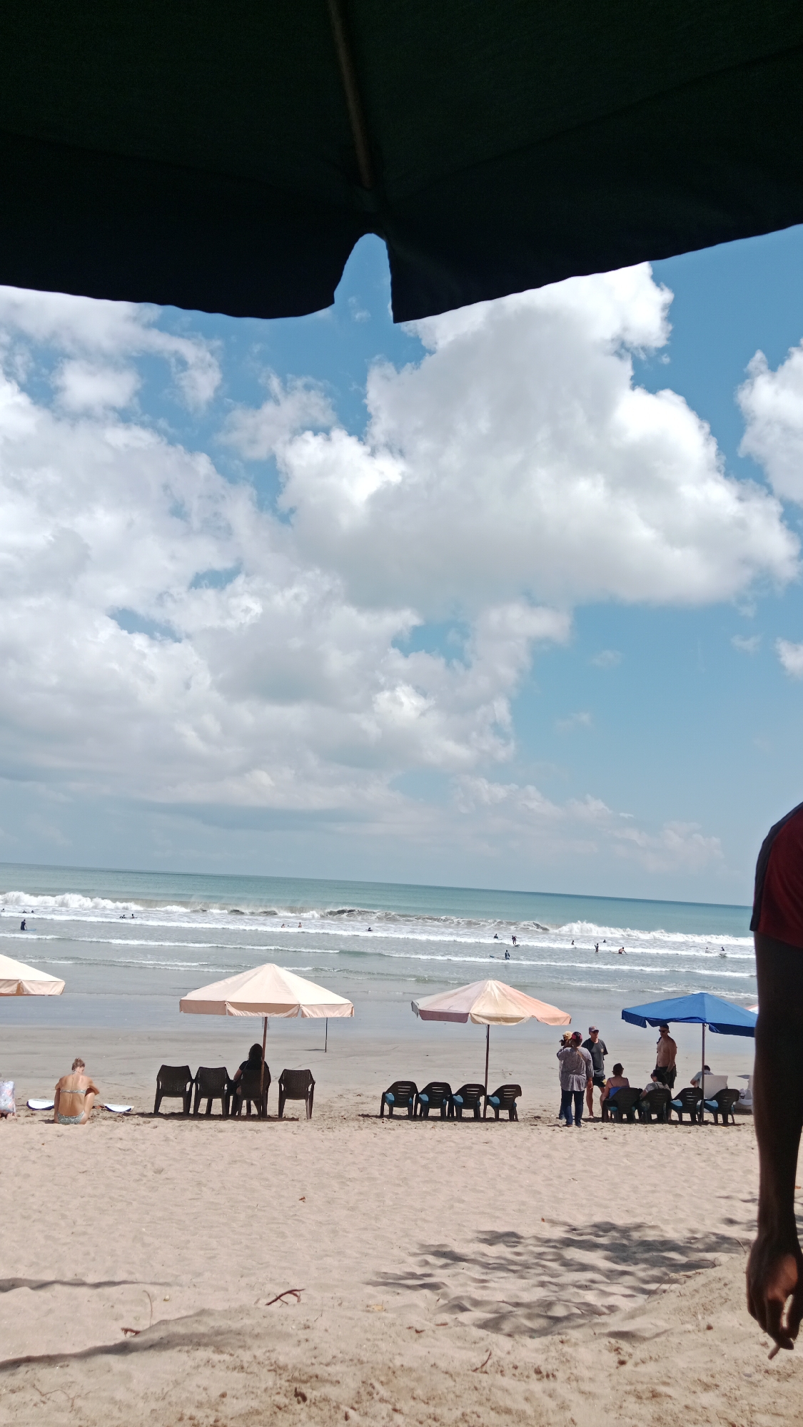 This image depicts a busy beach scene in Kuta, Indonesia, under a bright, partly cloudy sky. The foreground is dominated by the sandy beach, with the bottom edge of a large dark umbrella casting a shadow. Scattered across the sand are several beach chairs and umbrellas, some occupied and some empty.
In the middle ground, the ocean stretches out, with waves gently rolling towards the shore. Several people can be seen in the water, appearing to be swimming or surfing. Further back, the horizon is visible, where the blue of the sea meets the blue of the sky, dotted with white clouds.
In the foreground, on the left, a person in a bikini is seated on the sand, facing away from the camera. Towards the center, a group of people are gathered under an umbrella, some sitting in chairs, while others stand and converse. A person is holding a camera, potentially taking photos. On the right side, a person's arm, clad in a red shirt, is visible in the foreground, with their hand extended.
The scene conveys a lively atmosphere typical of a popular tourist destination like Kuta. The presence of umbrellas, chairs, and people enjoying the water suggests it is a well-utilized area for leisure and recreation. The overall impression is one of a warm, sunny day at the beach.