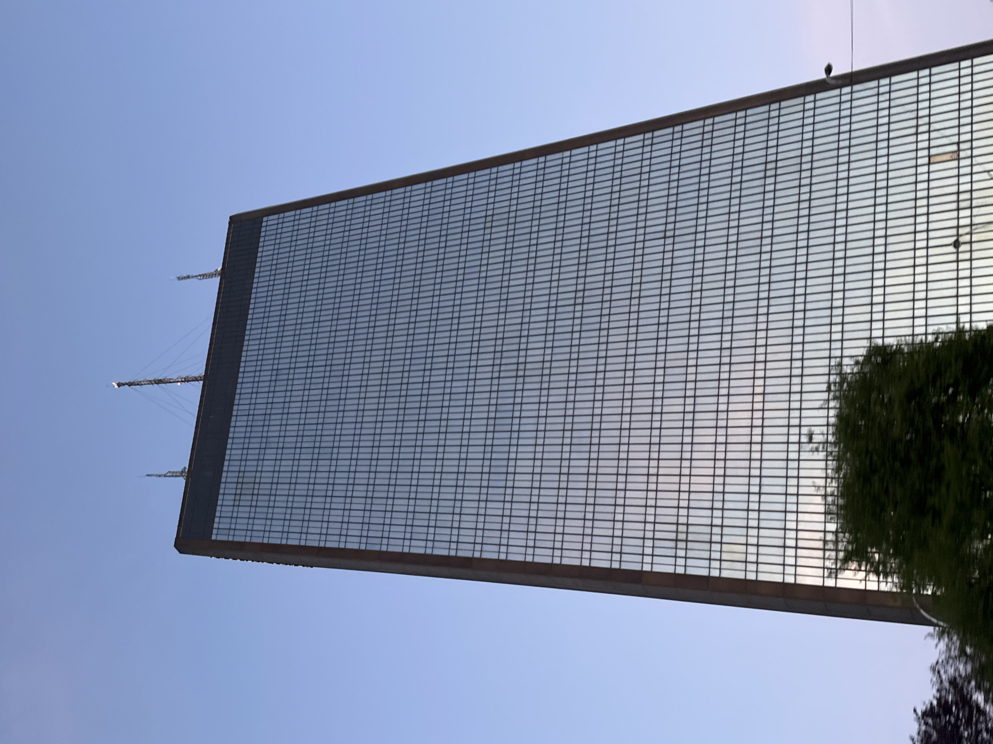 The image shows the Novotel Paris East, a skyscraper hotel located in Bagnolet, France. The building's facade features a gridded pattern of windows, reflecting the sky. The structure has several antenna on the roof and some nearby foliage. The sky is a pale blue, suggesting either early morning or late afternoon. The photograph is taken from a low angle, emphasizing the height of the building.