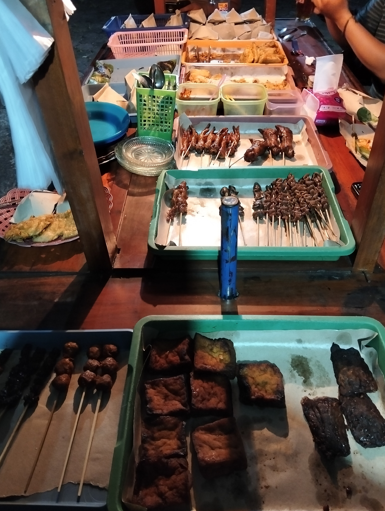 A food stall in Magelang, Indonesia, displays various Indonesian street foods on a wooden table. In the foreground, a green tray holds skewers of grilled tofu and small meatballs. To the left, another tray contains skewers of what appear to be grilled intestines and chicken feet. Further back, a blue plastic container and a green basket with utensils are visible. A stack of plates and a blue bowl sit on the table, alongside several paper-wrapped food packages. In the background, more food is presented in plastic containers, and a person's hands are visible holding a glass. The scene is lit with artificial light, casting shadows on the wooden surface.