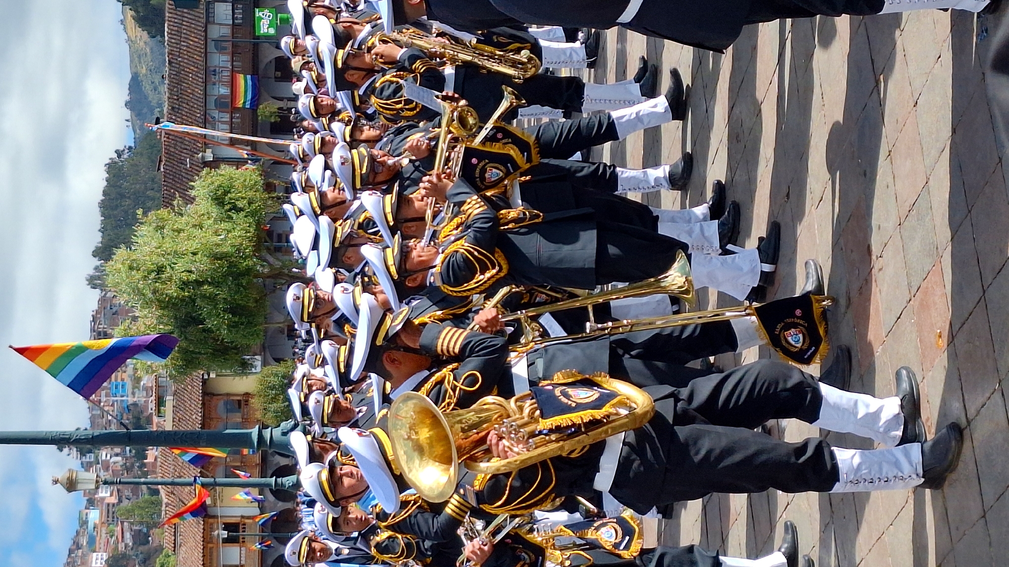 This image captures a marching band in uniform, likely participating in a parade or ceremony in the historic city of Cusco, Peru. The band members, predominantly young men, are dressed in dark navy jackets with gold trim and braiding, white shirts, white trousers with white gaiters, and white peaked caps with gold insignia. They are playing various brass instruments, including a large tuba, trumpets, trombones, and a saxophone, all of which appear to be made of polished brass. Many of the musicians are looking forward or at their instruments, suggesting they are in the process of playing or marching.
In the background, traditional Cusco architecture is visible, characterized by red-tiled roofs and the characteristic pastel-colored facades of buildings. A tall, dark lamppost stands prominently on the left side of the frame. Above the band, a rainbow flag is prominently displayed, its vibrant colors contrasting with the sky, which is partly cloudy. The ground is paved with interlocking cobblestones, suggesting the setting is a public square or street within the city. The overall atmosphere appears festive and organized, hinting at a cultural event or celebration in Cusco.
