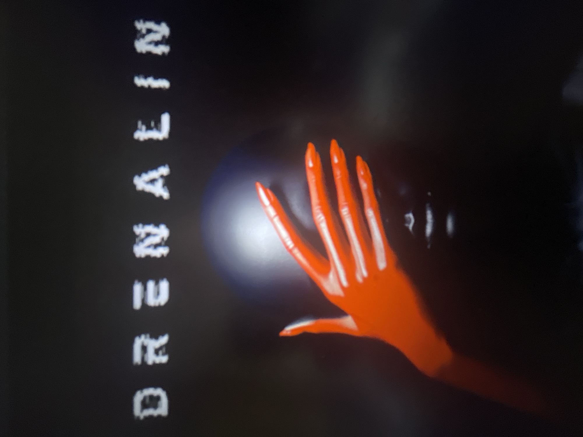 The image displays the word "DRENAALIN" in white, blocky, pixelated lettering across a dark, shadowy background. Below the text, a person's head is obscured by a dark, helmet-like object. A bright red, gloved hand with long, pointed fingernails is placed over the person's face, obscuring their eyes and nose. The hand appears to be a prominent focal point, with a glossy texture reflecting light. The overall mood is dark and somewhat mysterious. There is no information in the image to suggest a specific location in Punta Ala, Italy.