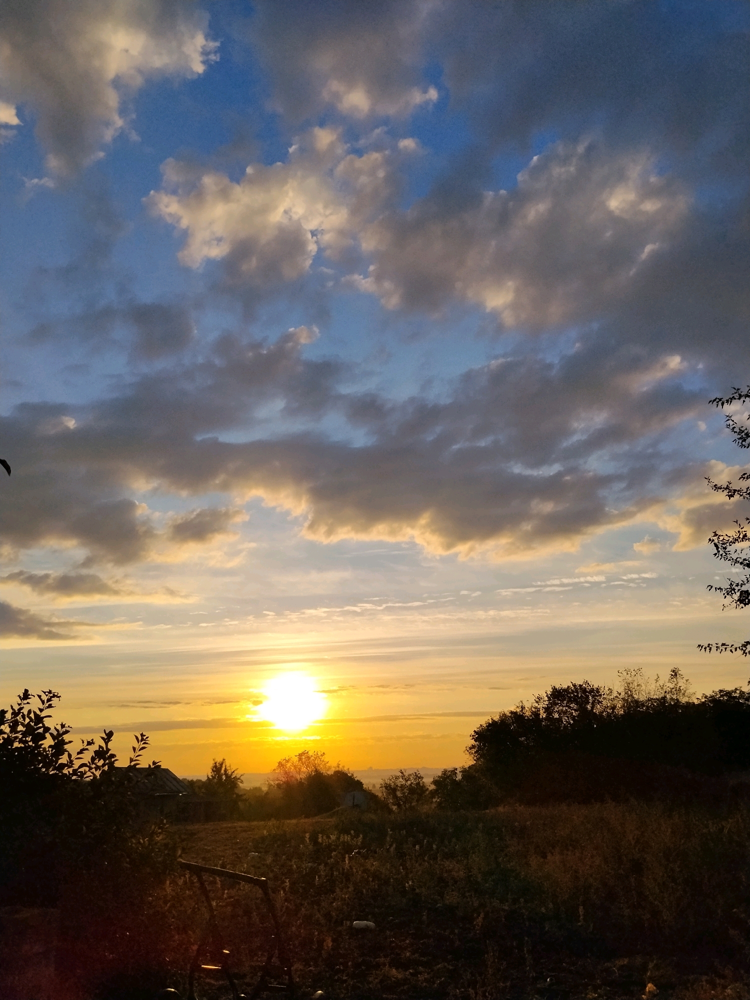 This is a photograph taken in Ustyvytsia, Ukraine, at either sunrise or sunset. The sky is filled with dramatic clouds, a mix of dark gray and lighter, golden-tinged formations, set against a bright blue backdrop. The sun, a brilliant orb, is partially obscured by clouds, casting a warm, golden light across the horizon.
In the foreground, silhouetted trees and foliage create a dark, textured border. A patch of tilled earth is visible, with some dry grass and a dark, metallic object that appears to be part of a tool or piece of equipment. In the middle ground, a small, rustic building with a partially collapsed roof is visible amongst the trees. The overall environment suggests a rural or semi-rural setting. There are no people visible in the image.