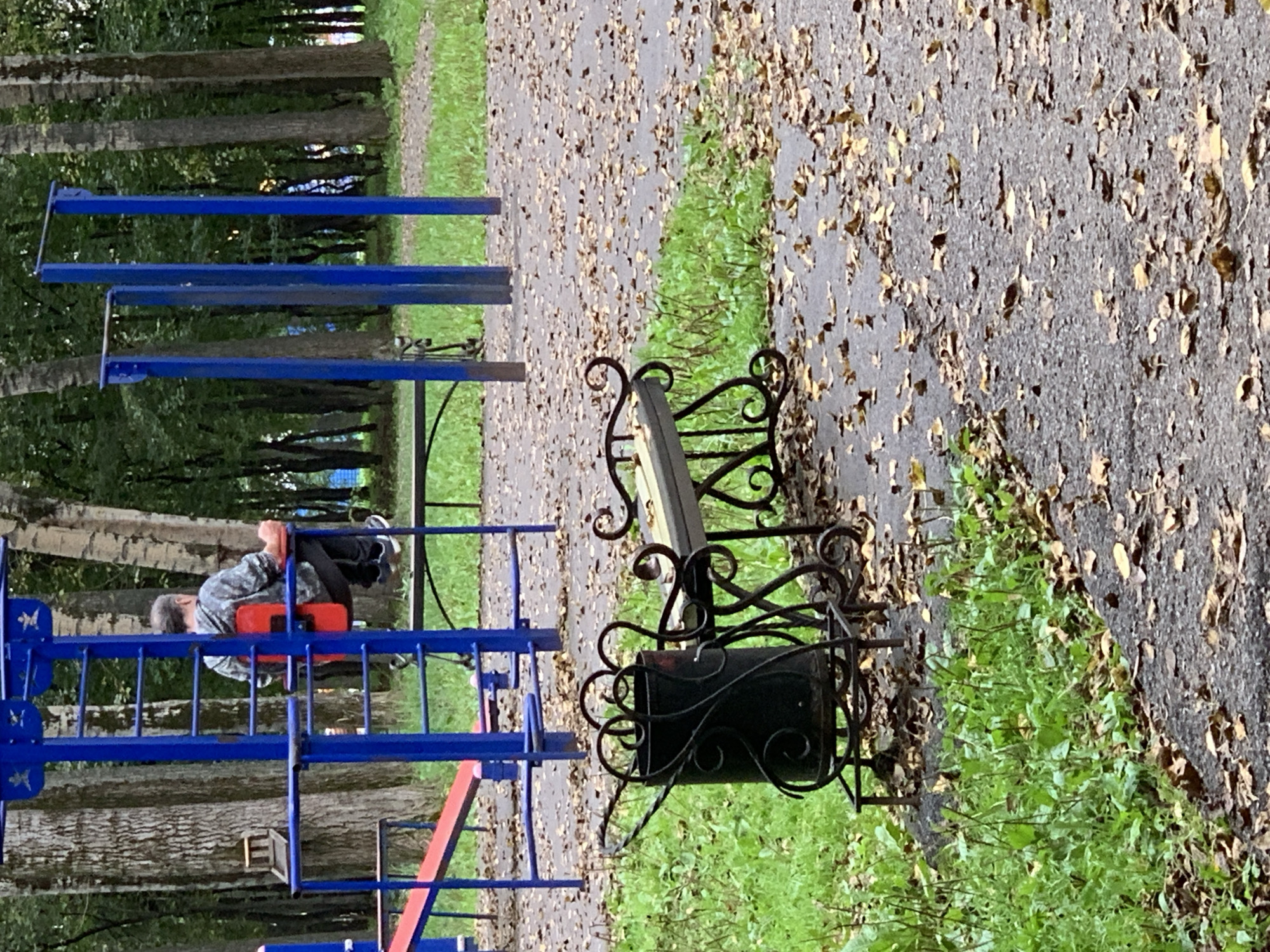 The image shows a man sitting on a red seat attached to a blue outdoor gym apparatus. He is wearing a grey hooded sweatshirt and black pants. The gym apparatus is located in a park, with trees visible in the background. There is a paved pathway covered in fallen leaves, and green grass on the side. In the foreground, there is an ornate black metal trash can and a matching bench. The scene appears to be in Prokopyevsk, Russia, a city known for its industrial history and located in the Kemerovo Oblast.