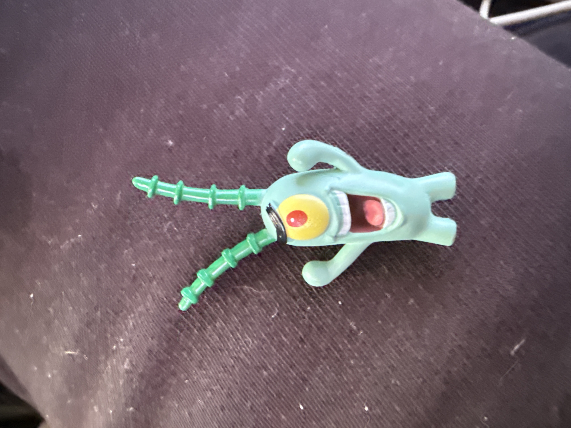 A light green plastic toy, resembling the character Plankton from SpongeBob SquarePants, is lying on a dark purple fabric surface. The toy has two green antennae with segmented details, a single large eye with a yellow iris and red pupil surrounded by a black band, and a wide open mouth revealing pink interior and white teeth. Its arms are outstretched. The fabric surface appears to be clothing, possibly pants or a jacket, and is covered in small white specks, likely lint or dust. The lighting suggests an indoor environment. No other subjects or discernible elements of a city or specific location are present.