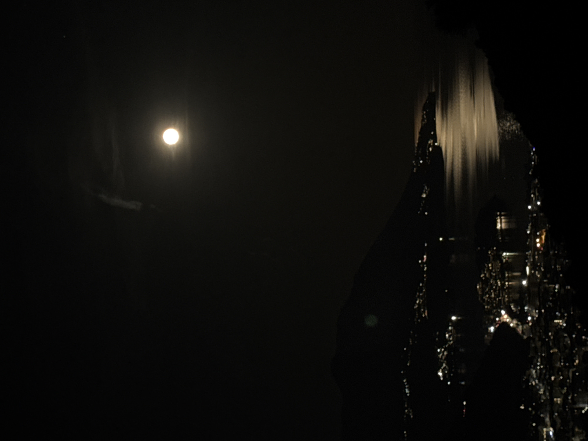 The image depicts a night scene in what appears to be a city. A bright moon is visible in the upper left of the frame. The right side of the image features a dark, jagged shape that could be a mountain or rock formation, with sparse light reflecting off of it. A bright waterfall is visible to the right of the formation. The lights appear to be city lights reflected on the water's surface, implying that the city is located near a body of water. Given the context, this could be in the vicinity of Podostrog, Montenegro. The overall impression is one of darkness and sparse illumination.