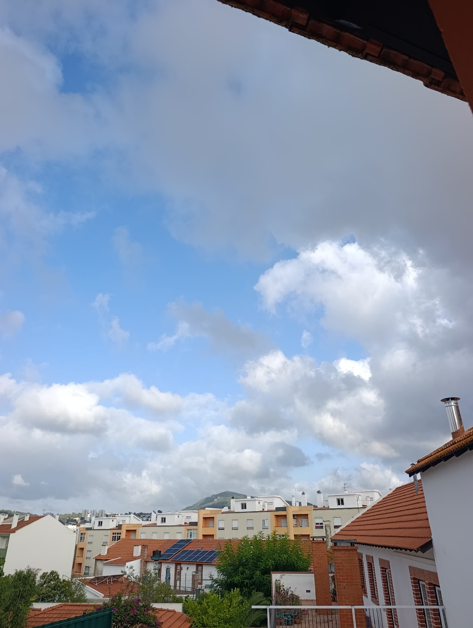 The image shows a view of a city with buildings and a sky filled with clouds, likely taken from a balcony or rooftop in Setúbal, Portugal. The foreground is dominated by the edge of a building with a terracotta tiled roof and a visible chimney. Below, several other buildings are visible, some with white facades and terracotta roofs, and others with orange and gray exteriors. Some of these buildings have solar panels installed on their roofs. Lush green trees and foliage are scattered between the buildings. In the background, a range of hills or mountains can be seen, with more buildings and structures at their base. The sky is a mixture of bright blue patches and large, fluffy white and gray clouds, suggesting an overcast or partly cloudy day. There are no people visible in the image. The scene depicts a residential or urban environment.