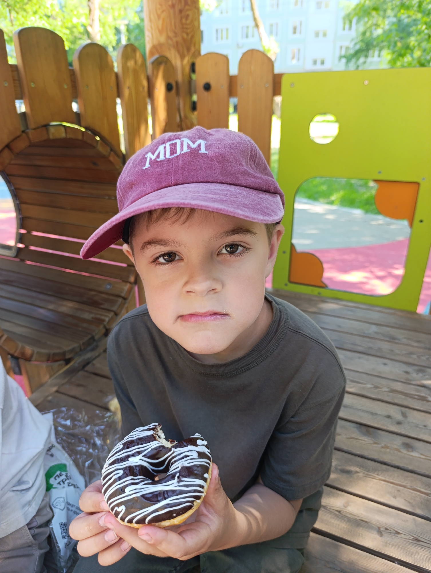 An outdoor scene captures a young boy, approximately 6-8 years old, sitting on a wooden slatted deck, likely part of a playground. He wears a faded purplish-pink baseball cap with the word "MOM" embroidered in white uppercase letters on the front, and a dark olive green short-sleeved t-shirt. The boy has dark eyes and a neutral to slightly serious expression with a subtle downturn to his lips, as he looks directly into the camera. He holds a partially eaten round donut with dark chocolate frosting and white zigzagging icing in both hands. To the boy's left, a transparent plastic bag containing a green logo and partially visible white Cyrillic text (suggesting a Russian retail chain, such as Pyaterochka) rests near what appears to be another person's light-colored pants or garment. The background features a wooden playground structure resembling a half-barrel or tunnel, and a modern bright lime green rectangular playground panel with rounded corners, a circular cutout, and an orange semicircular cutout. Beyond these structures, green foliage from trees is visible, and in the distant background, a multi-story light blue residential building with numerous windows, characteristic of Soviet-era or post-Soviet architecture, confirms the location as Moscow, Russia. The bright daylight indicates a clear day.