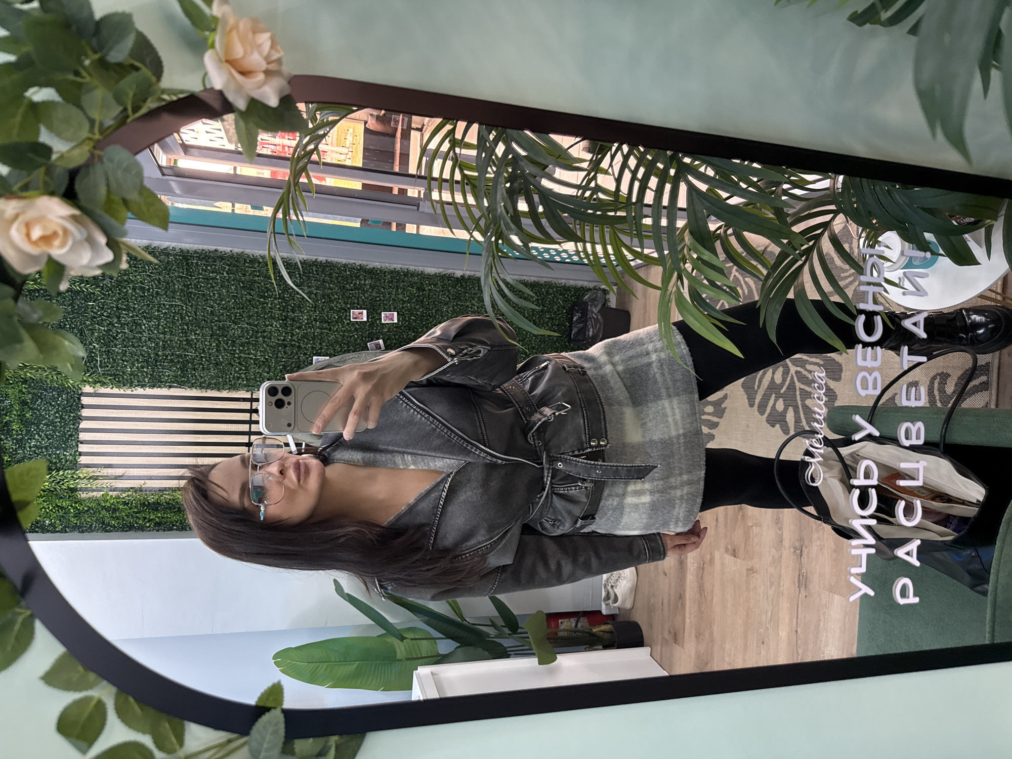A woman with long brown hair and glasses takes a selfie in a decorative mirror. The mirror is adorned with artificial flowers and greenery. The woman wears a gray jacket and dark leggings. The floor is light wood-colored. The mirror reflects an interior with a plant, a green wall, and a view out a window to a building. The Cyrillic text "Учись у весны расцветать!" (Learn from spring to bloom!) is partially visible in the lower right of the mirror. The photo was taken at, or near, Chaikovsky, Perm Krai, Russia.