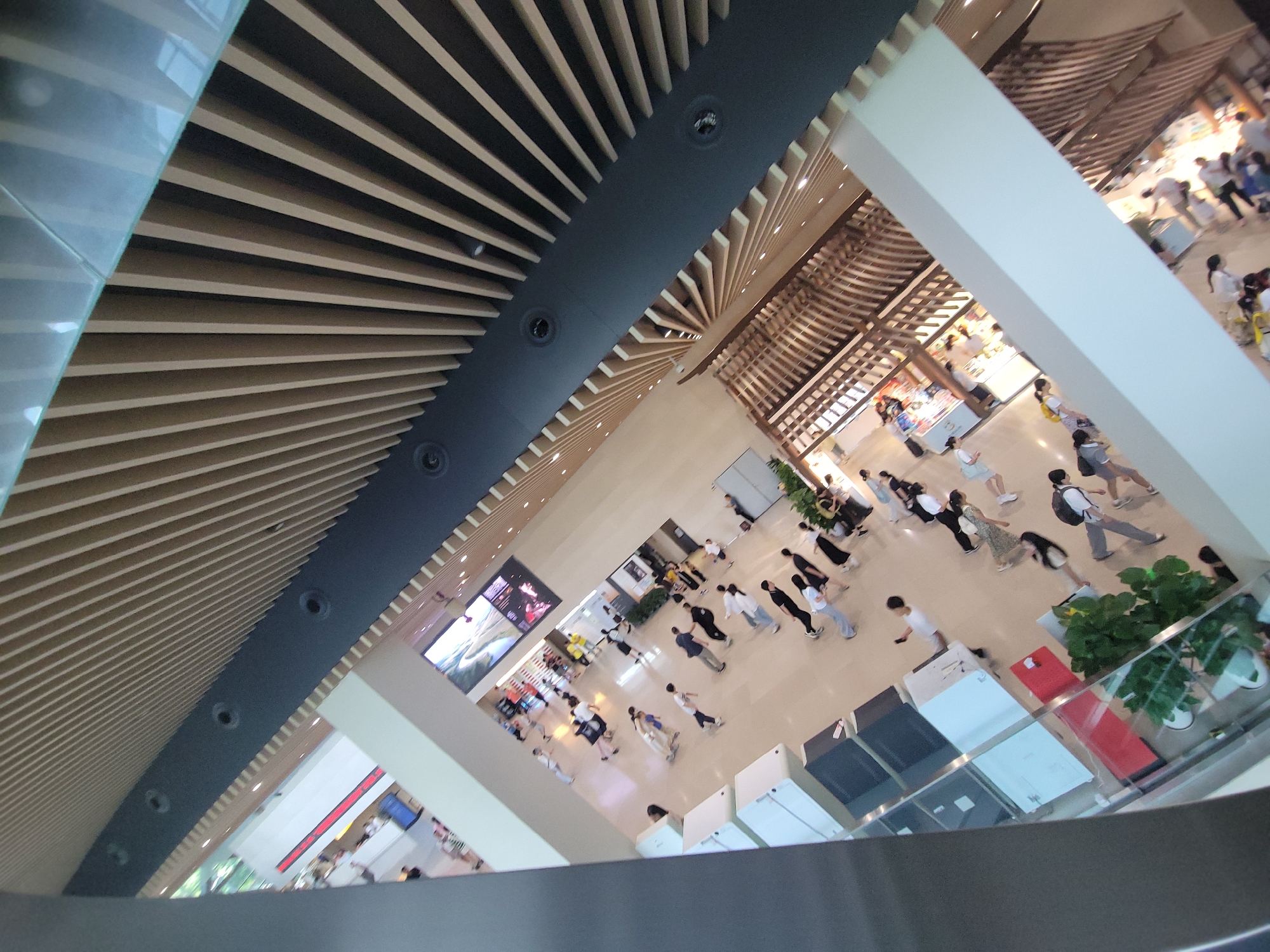 The image captures an aerial view of a bustling interior space, likely a shopping mall or public atrium, in the Qingyang District of China. The scene is characterized by a high, slatted wooden ceiling with dark architectural elements and circular lights. Below, numerous people are spread across a wide, light-colored floor. Many individuals appear to be walking, with some in groups and others alone. Several people are dressed casually in t-shirts, shorts, and pants, and some are carrying bags. The environment includes large display screens, store facades with merchandise visible, and modern architectural features like white pillars and glass railings. Green potted plants are also interspersed throughout the space. The image is taken from a high vantage point, looking down at an angle, which gives a sense of scale and activity.
