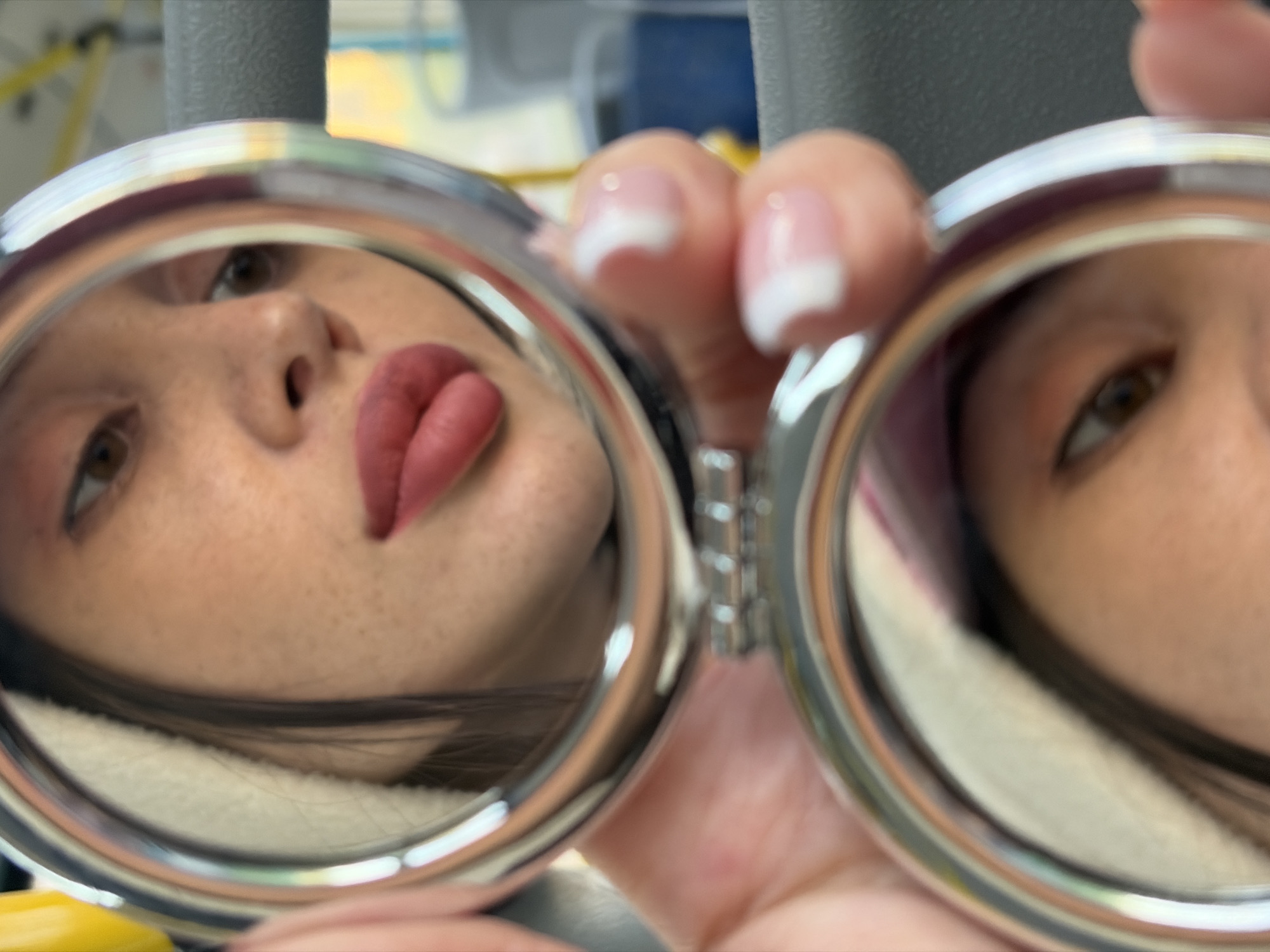 A person holds a double compact mirror. The left mirror reflects a close-up of their face showing their nose, lips painted red, and left eye. The right mirror reflects a closer view of the same person's right eye. The person's fingers with white-tipped nails hold the compact mirror. The background is out of focus, showing a yellow object and gray poles. The location is not discernible from the image.