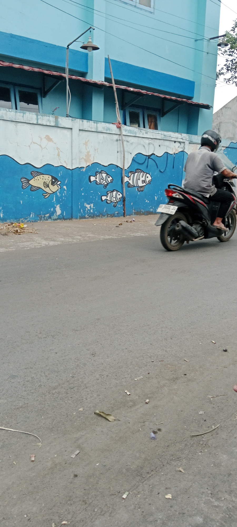 A man wearing a grey t-shirt and helmet rides a dark-colored motorcycle to the right on a paved road. The road is asphalt and appears to be in a moderately urban or suburban area, with debris scattered on the surface. To the left of the road, a light blue building is visible, with a light blue accent band across its facade. Below the building, a light grey wall features a mural of blue wavy water with stylized fish. One large yellow fish with black markings is on the left, and three smaller black and white striped fish are to its right. A streetlamp hangs from a pole in front of the building.