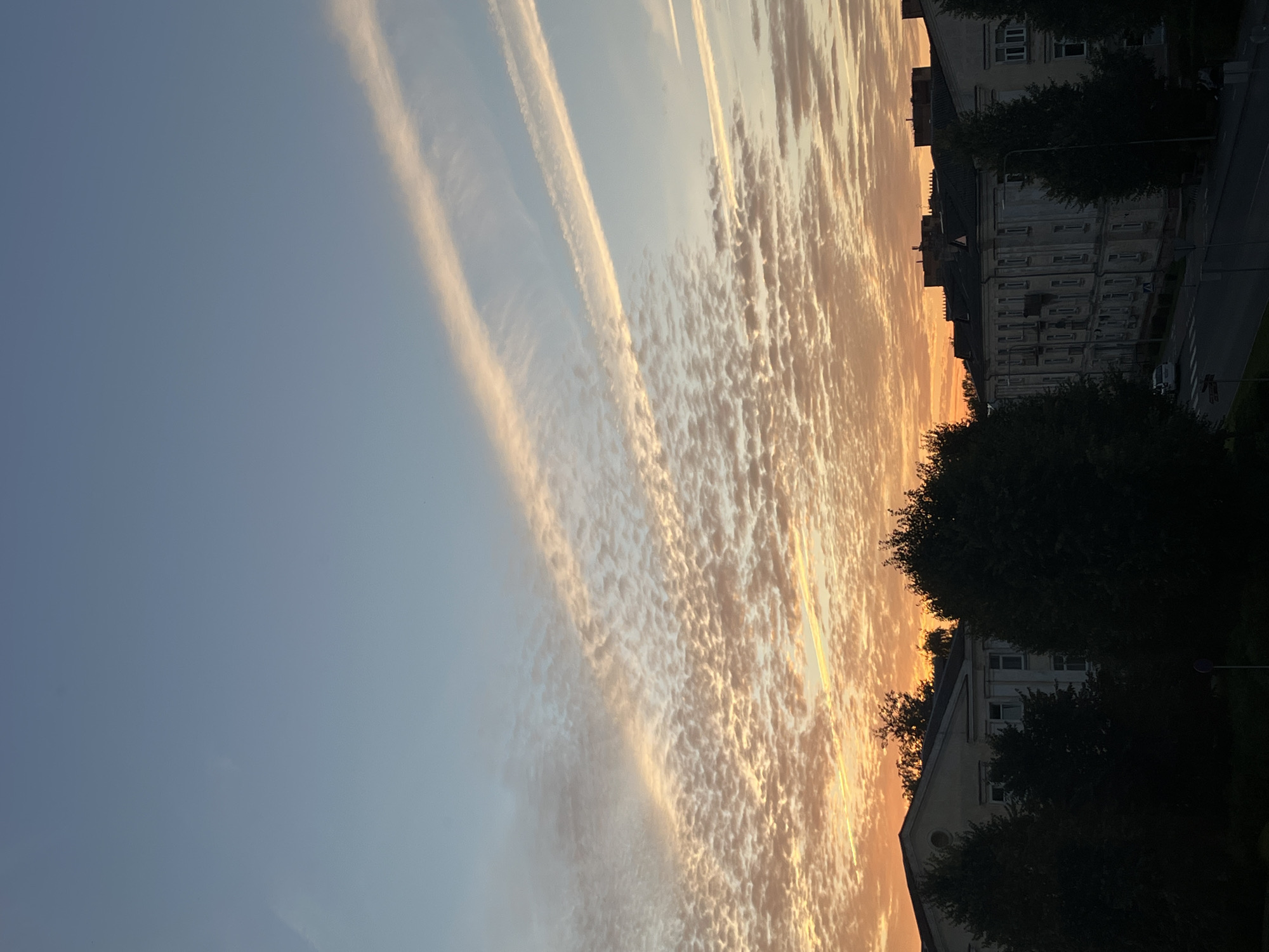 This is a vertical, eye-level, outdoor, handheld shot of a city skyline during a sunset. The sky dominates the upper portion of the frame, transitioning from a pale blue at the top to an orange and pink gradient closer to the horizon. The sky is filled with a textured pattern of small, wispy clouds, with two distinct, parallel contrails streaking across the upper right section. Below the sky, silhouetted against the bright horizon, are the dark shapes of trees and buildings. The buildings appear to be multi-story apartment blocks with light-colored facades and dark roofs. A street runs horizontally across the lower portion of the image, with a single white car visible on it. The general impression is one of a peaceful urban evening scene.