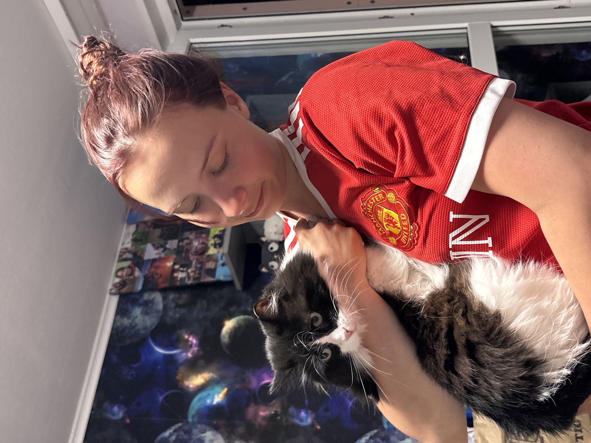 A woman is holding a black and white cat. The woman has auburn hair styled in a bun and is wearing a red shirt with white accents. The cat is held close to her chest. The background includes a window and a wall with a space-themed design. There is also a collage of images on the wall. The shirt has a logo with the word Manchester. The shirt also has a part of the word "UNITED". The image was taken indoors.