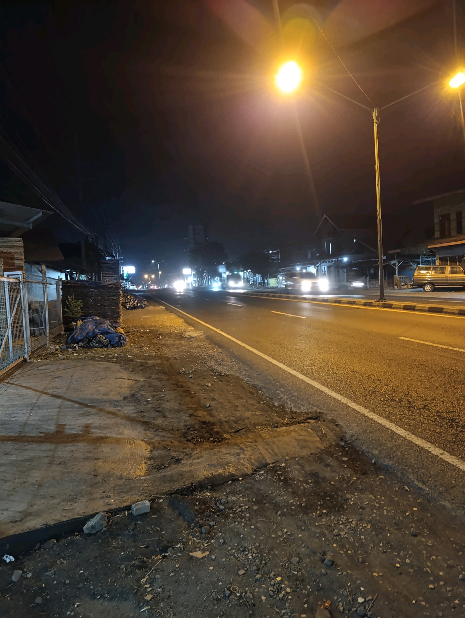 The image captures a nighttime street scene in Karangsono, Indonesia. A paved road with a white lane divider occupies the right side of the frame, with streetlights casting a warm glow and creating lens flares. Several vehicles, including what appears to be a minivan on the right and other cars with illuminated headlights, are traveling along the road. The left side of the street features a sidewalk area with a wire fence bordering a building or structure. Piles of lumber and a large blue tarp covering debris are visible along the roadside. The environment is dimly lit, typical of a street at night.