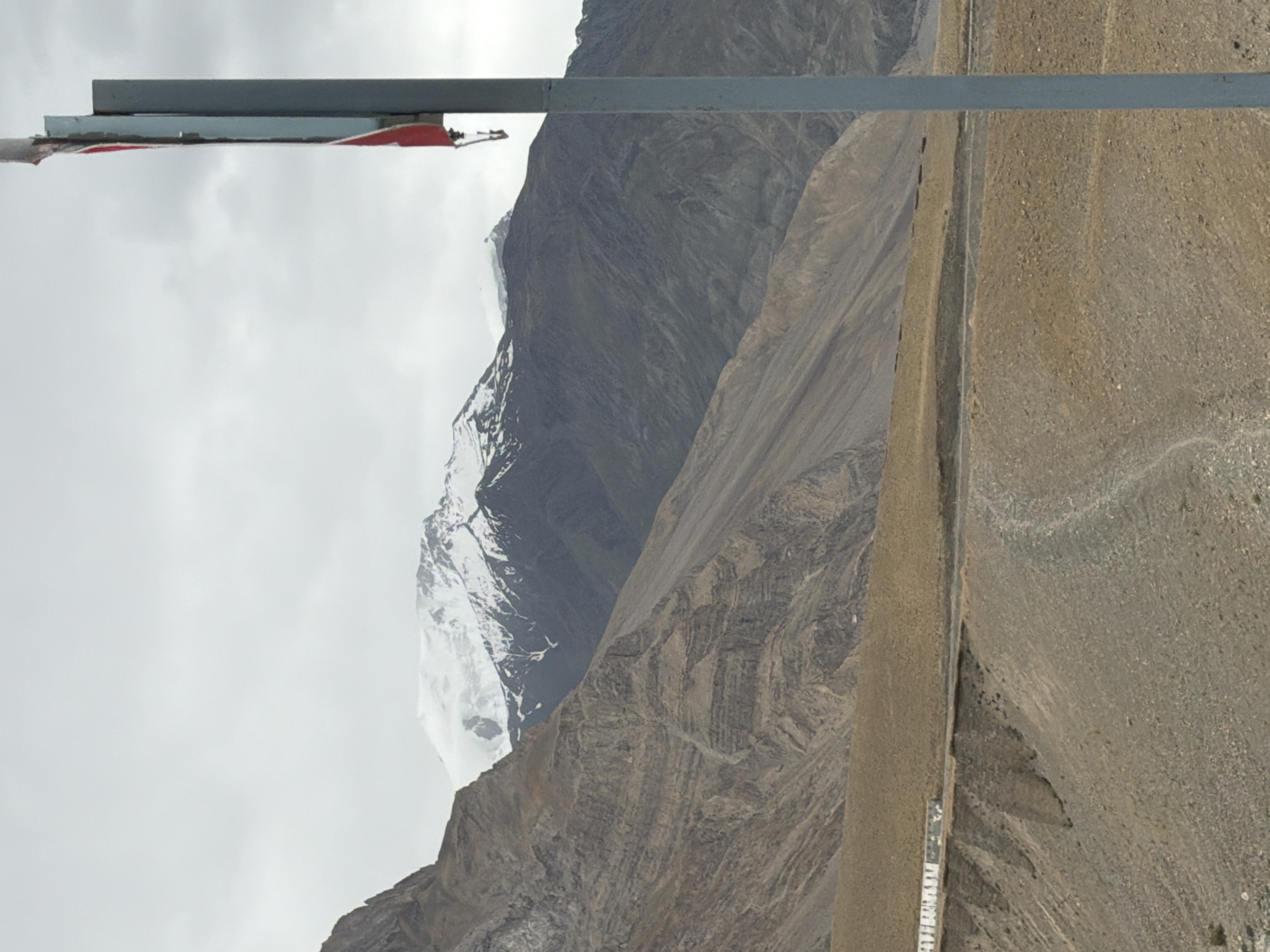 A vertical, full-shot image depicts a mountainous landscape under a cloudy sky. In the foreground, a tall, gray pole with a partially visible red and white sign is positioned on the right side. The terrain slopes upwards from the foreground to midground, featuring dry, rocky ground and a winding dirt road. A low, white structure with a patterned camouflage-like front runs along the edge of the road in the midground. Beyond this, imposing, layered mountains rise, their sides textured with erosion. The highest peaks in the background are capped with snow and partially obscured by clouds. The scene is situated in Zangla, India, a remote village in the Himalayas known for its stark, beautiful terrain.