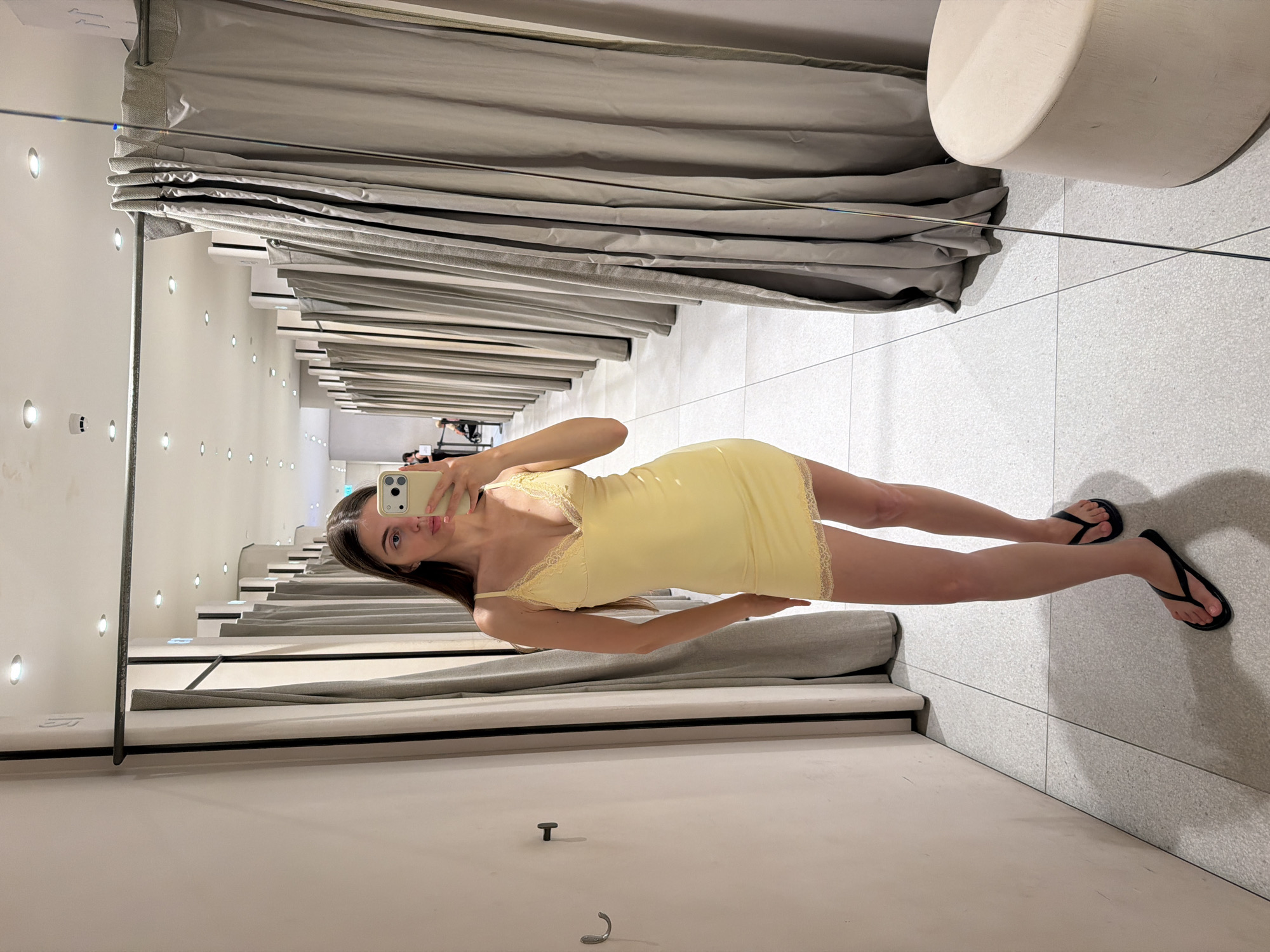 A woman stands in a fitting room, taking a mirror selfie with her phone. She is wearing a yellow slip dress with lace trim and black flip-flops. The fitting room has light-colored walls and floor, with gray curtains hanging on either side. A metal rod runs across the top of the fitting room, supporting the curtains. In the background, several other fitting rooms are visible, along with a stool. The overall setting suggests a retail environment.