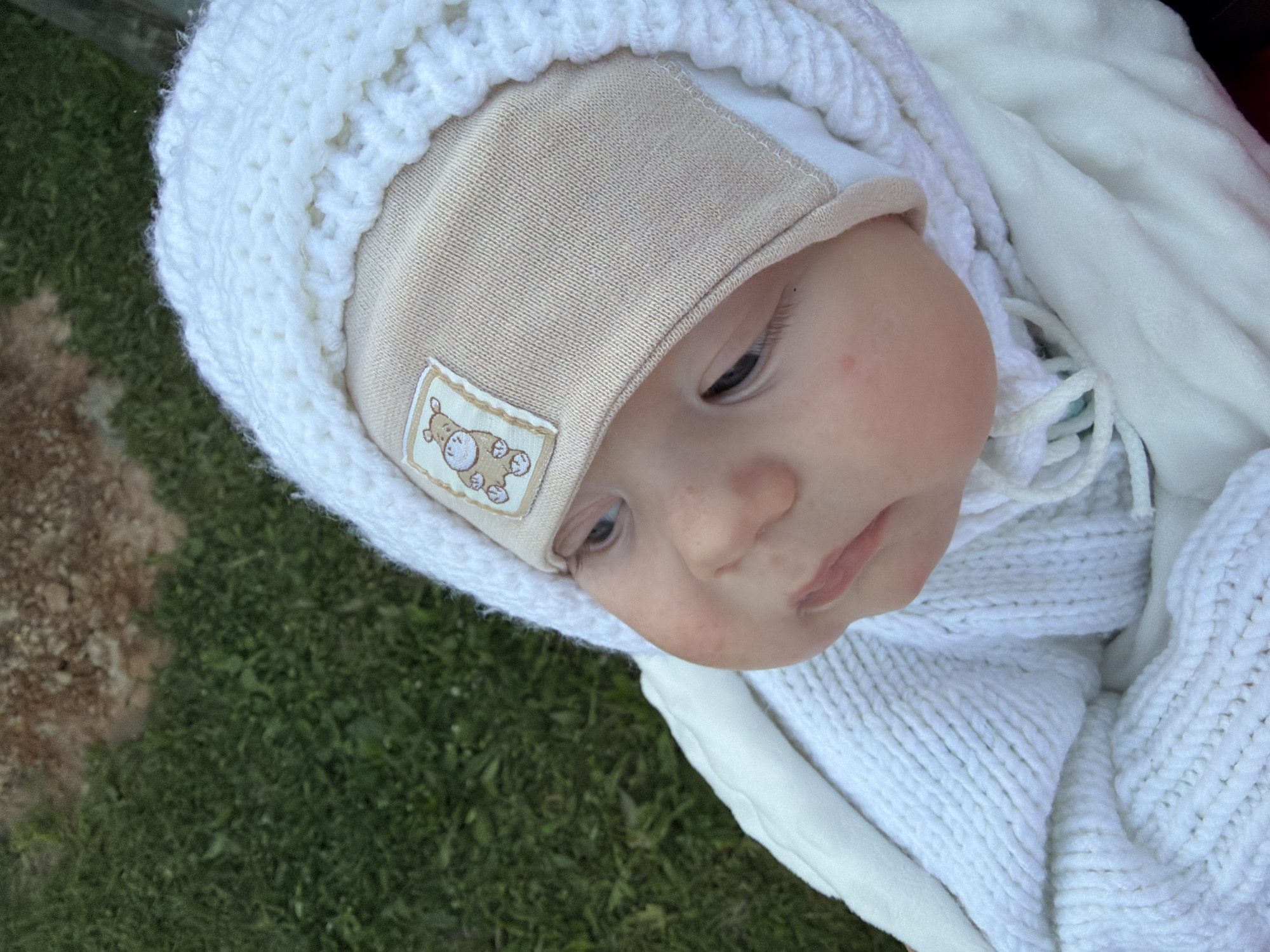 A close-up, high-angle, outdoor shot shows the face of a baby wrapped in white knitted clothing and a beige hat with a small patch of a cartoon hippopotamus. The baby's face is turned to the left, with its eyes looking downwards. The background consists of green grass and some brown soil. The image was taken in the village of Krasny Mayak, Russia.