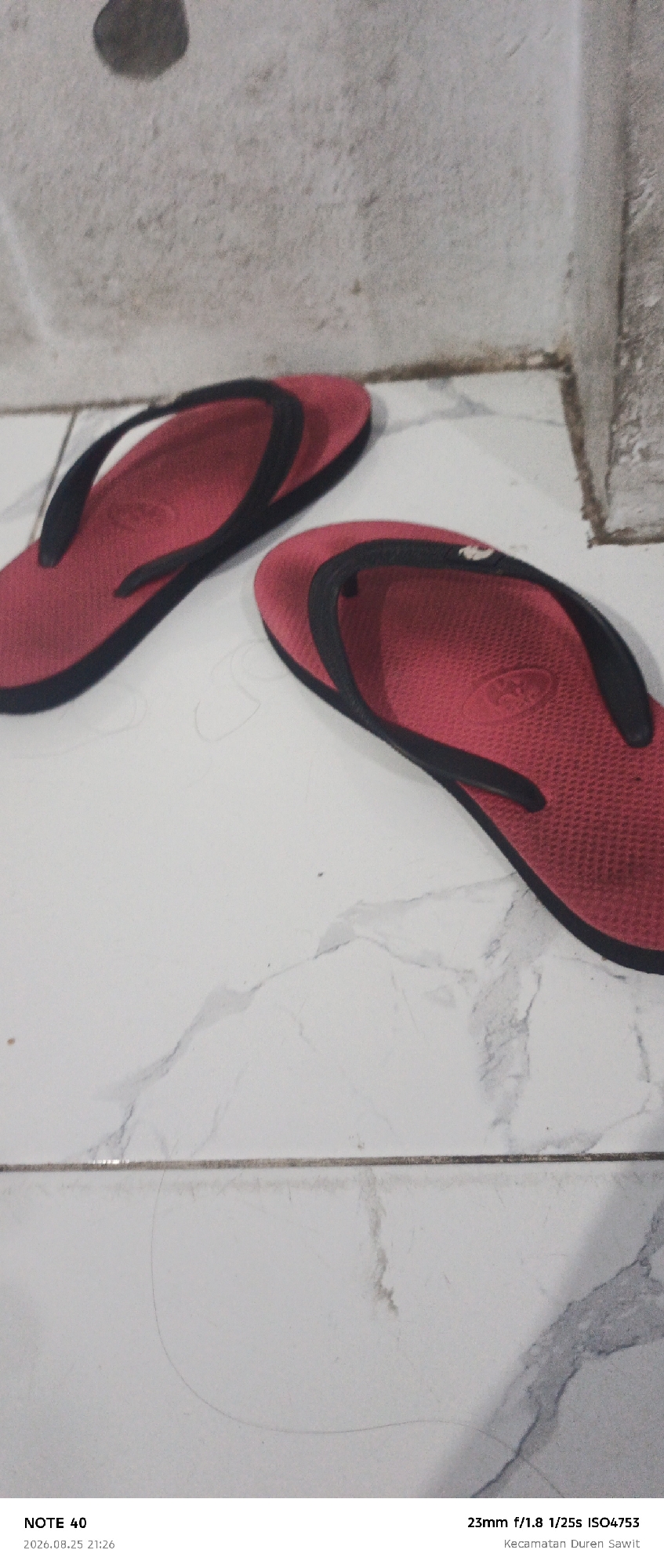 A pair of red flip-flops with black straps are displayed on a white tiled floor. The left flip-flop is positioned horizontally and angled slightly upwards towards the left. The right flip-flop is placed to the right of the first and is angled downwards towards the right. The flip-flops are in the foreground, and the background consists of a textured light-colored wall. The scene is well-lit, with some shadows cast by the flip-flops. The image is a close-up shot, emphasizing the details of the footwear and the surrounding environment. The text overlay indicates that the photo was taken with a "NOTE 40" device, on "2026.08.25 21:26" with camera settings "23mm f/1.8 1/25s ISO4753", and a location identified as "Kecamatan Duren Sawit", which is a subdistrict in East Jakarta, Indonesia.