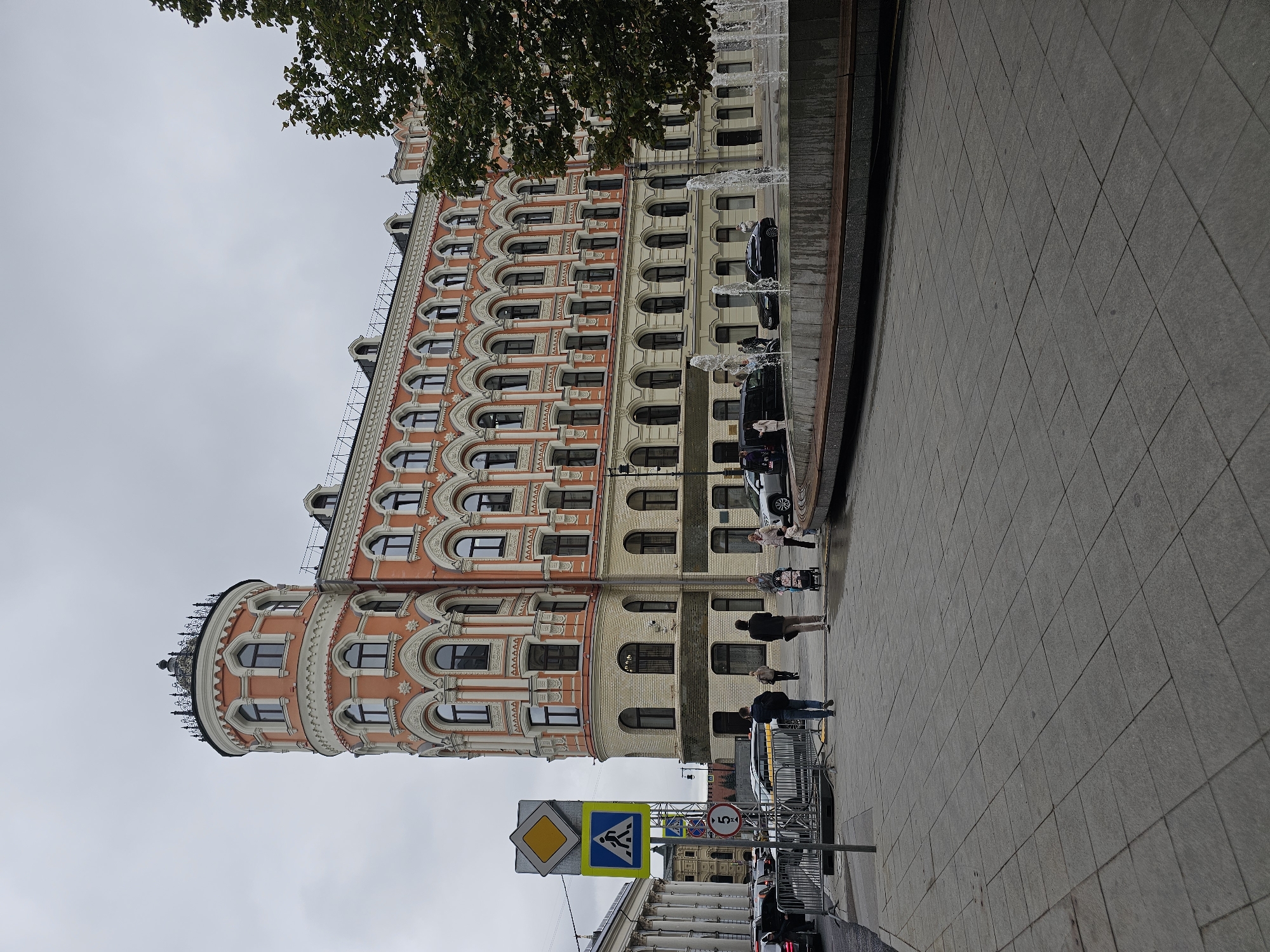 The image depicts a street scene in Moscow, Russia, featuring a prominent, ornate building with salmon-colored walls and cream-colored architectural details. The building has a rounded corner section topped with a decorative cupola. In front of the building, a sidewalk area with large square tiles is visible, leading to a fountain with multiple jets of water.
Several people are present in the scene. Two individuals are walking away from the camera on the sidewalk, and a woman is pushing a stroller. Further down the street, more pedestrians can be seen. Several cars are parked or driving on the road in the background. Traffic signs are also visible, including a pedestrian crossing sign and a speed limit sign. The sky is overcast, suggesting cloudy weather. The overall atmosphere appears to be a typical urban environment in Moscow.