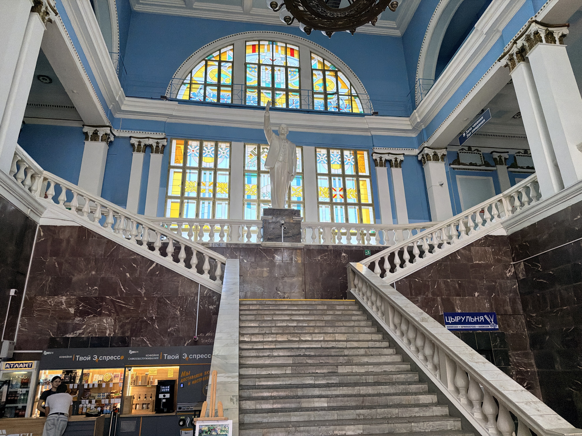 The image depicts the interior of the Homyel railway station in Homyel, Belarus. A grand staircase with marble cladding and ornate white balustrades dominates the foreground, ascending towards a large, arched stained-glass window. The window features a vibrant pattern of yellow, blue, and red glass. In front of the window stands a white statue of a man with his arm raised. The walls are painted a striking blue, accented with white molding and columns topped with gold detailing. Below the staircase, a cafe or shop with signage reading "Твой Эспресс" (Your Express) is visible, with two individuals standing at the counter. To the right of the staircase, a blue sign with white lettering reads "ЦЫРУЛЬНЯ" (Hairdresser). The overall scene conveys a sense of civic grandeur and functionality, characteristic of a major transportation hub.