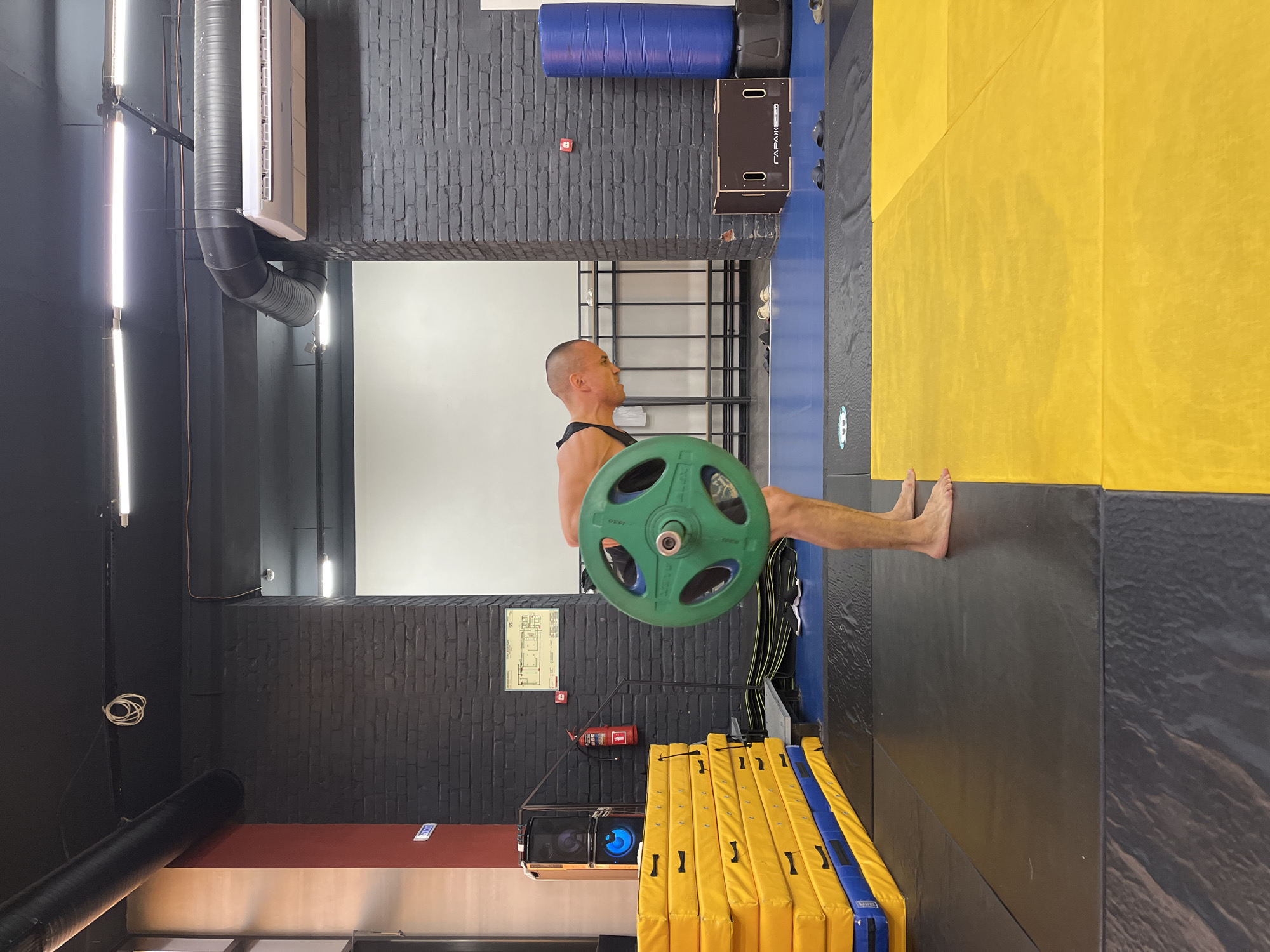 A bald man is in a gym, performing a deadlift with a green weight plate. He is bending over with a straight back, holding the barbell. The gym has black brick walls, industrial piping, and fluorescent lighting. There are stacks of yellow and blue mats, gym equipment like a punching bag, and a fire extinguisher visible. The image appears to be taken in Krasnoyarsk, Russia.