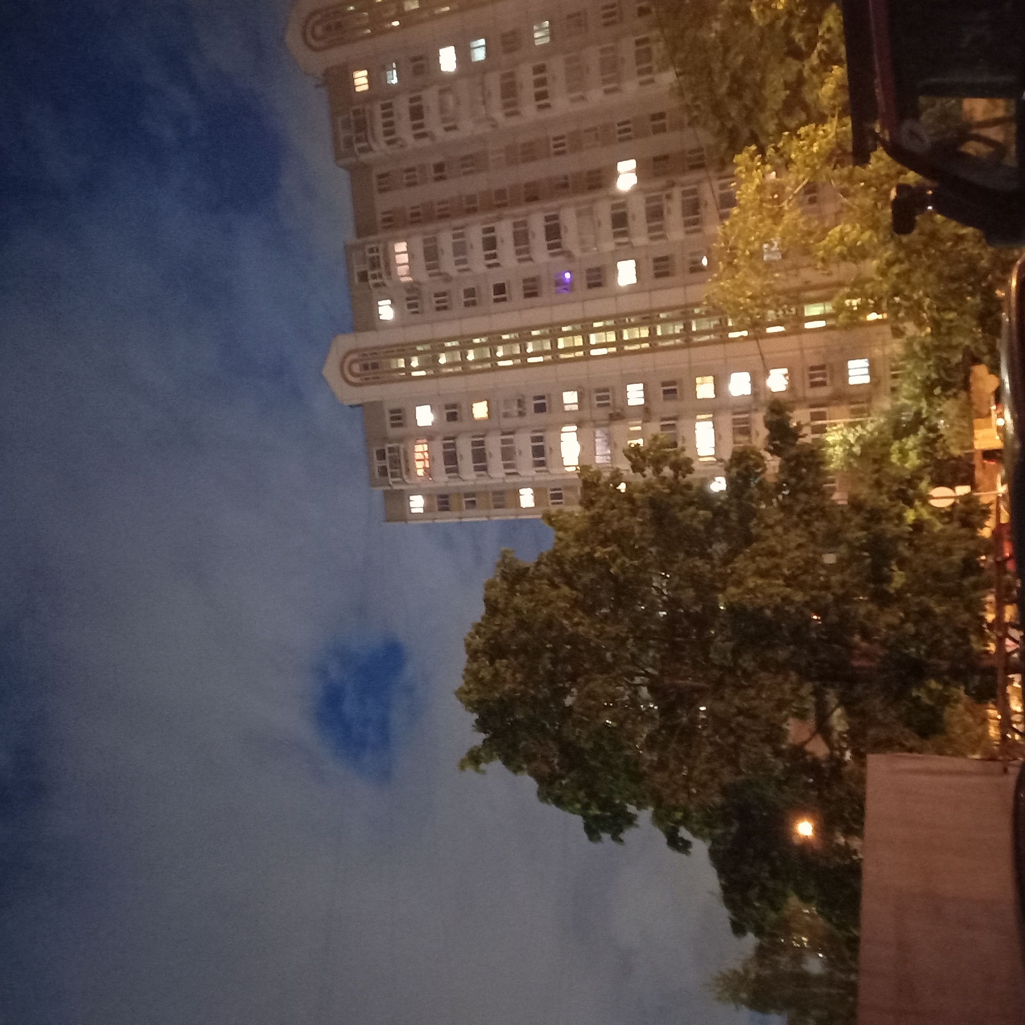 The image, taken at night, depicts a tall building, possibly residential, with multiple illuminated windows, partially obscured by foliage from trees on the right side. The sky, which takes up a significant portion of the image, is a mix of dark blue and cloudy patches. The composition is slightly rotated, giving a tilted perspective of the building. The building's architecture suggests a contemporary style, with a repetitive pattern of windows and balconies. There is a streetlight visible in the bottom-right corner. Given the style of architecture and the presence of dense urban foliage, it's possible that the location is in a developed residential area in the city of Moscow, Russia.