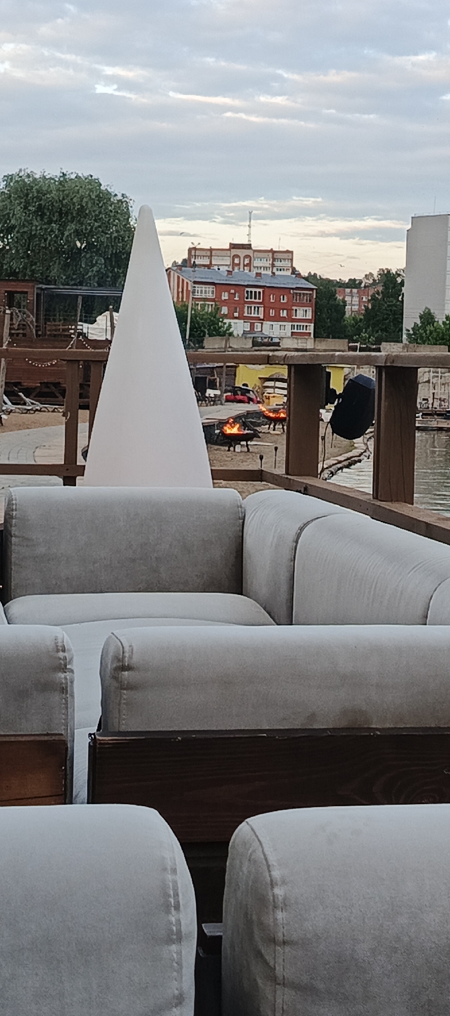 The image depicts an outdoor seating area with a view of buildings across a body of water, likely in Votkinsk, Russia. In the foreground, there are light-colored upholstered sofas and wooden furniture. A large, white cone-shaped object stands prominently in the mid-ground. Beyond the seating area, several fire pits with flames are visible, suggesting a gathering or entertainment space. In the background, red-brick buildings with multiple windows are situated along the water's edge, and a taller white building is also present. Green trees are interspersed with the structures, and the sky is partly cloudy. The overall scene appears to be a waterfront establishment, possibly a cafe, restaurant, or recreational area.