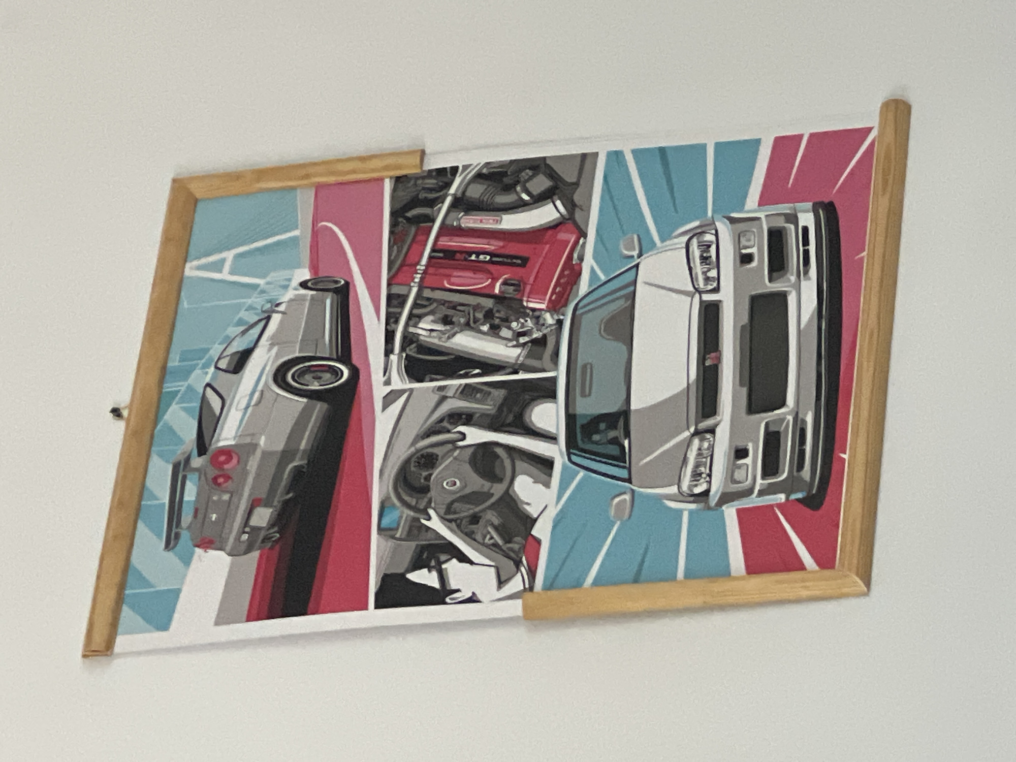 The image shows a framed poster hanging on a white wall. The poster is divided into three horizontal panels, each featuring an illustration of a car. The top panel displays the rear view of a silver Nissan Skyline GT-R. The middle panel shows a close-up of a person's hands on a steering wheel inside a car, and a view of a red engine. The bottom panel shows the front view of a silver car, likely another Nissan Skyline GT-R. The poster has a wooden frame and is hung with what appears to be a string. The lighting suggests an indoor environment. There is no identifiable context within the image that would place it in Cluj-Napoca, Romania.