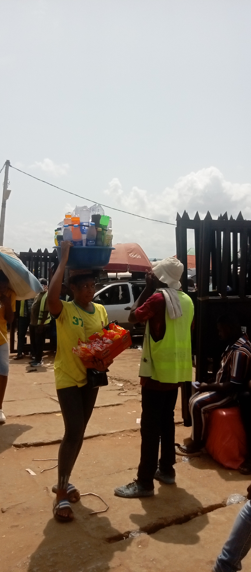 Onitsha, Nigeria. A woman carrying a tray of bottled drinks on her head and a box of snacks in her hand stands on a dusty, light-colored surface, apparently negotiating a transaction with a man wearing a yellow safety vest and a light-colored hat. A fence, a car, and other people are in the background.