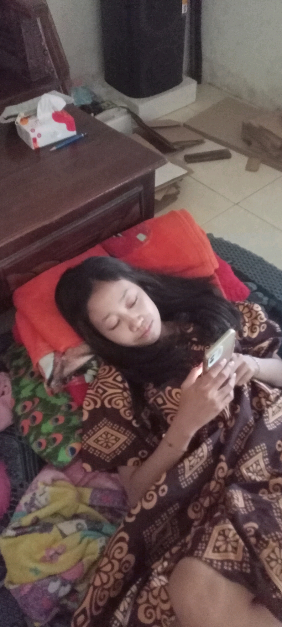 The image shows a young person lying down, holding a smartphone. They are wearing a brown and tan patterned garment. The person is resting on a surface covered with blankets and pillows, one of which is orange. To the left of the person, there is a wooden desk with a tissue box and a pen on it. In the background, a large black cylindrical object is visible. The floor appears to be tiled. The overall impression is one of a relaxed, indoor setting. The image appears to be taken in Gempolklutuk, Indonesia.
