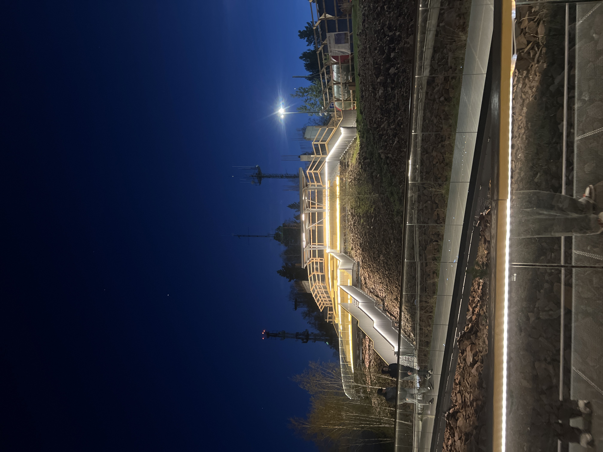 The image shows a lit staircase in the city of Krasnoyarsk, Russia at night. The dark blue sky is visible. There are radio towers in the background. The staircase is well-lit and appears to be of modern design with glass panels and wooden accents. The surrounding terrain is rocky and uneven. People are walking on the stairs.