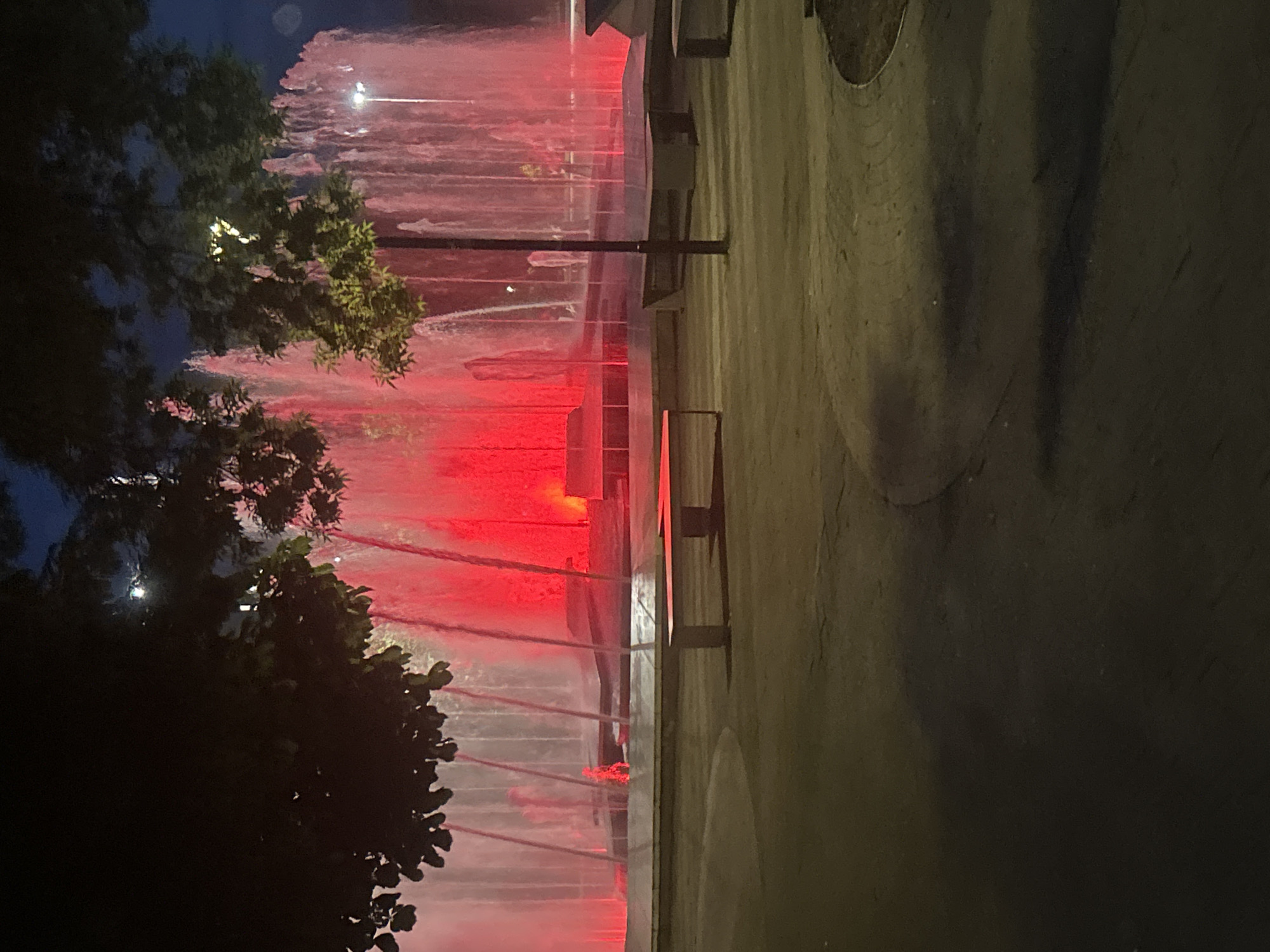 The image depicts a fountain in Pleven, Bulgaria, at night. The water jets are illuminated with red lighting. Trees are visible on the left side of the frame. To the right of the fountain is a paved area with benches. The scene is dark, with limited ambient light.