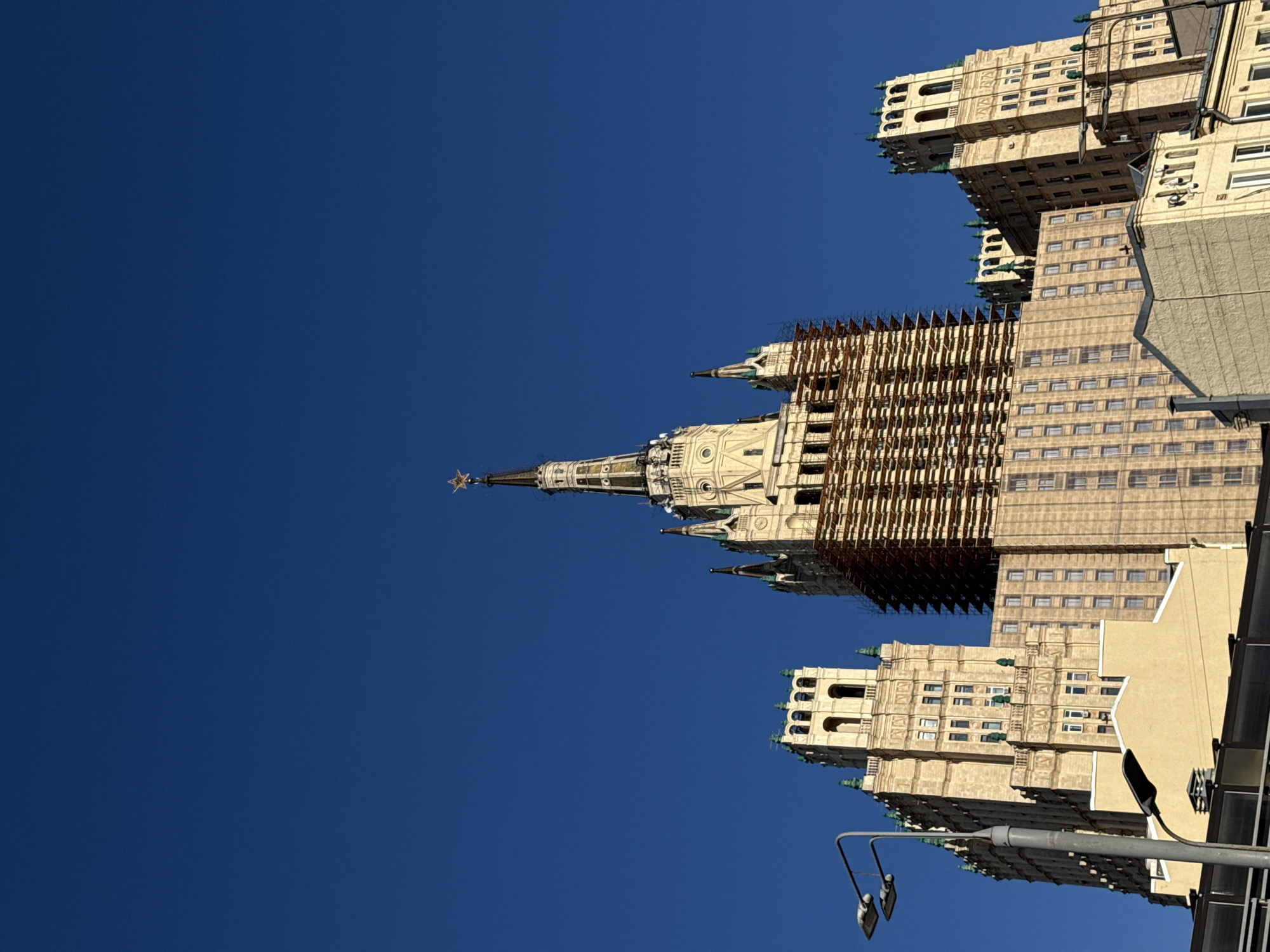 The image shows a low-angle, close-up shot of the Kotelnicheskaya Embankment Building in Moscow, Russia, under a clear, bright blue sky. The building, a prominent Stalinist skyscraper, is characterized by its ornate architecture, featuring multiple towers and decorative elements. The central spire, crowned with a red star, is a focal point. Scaffolding is visible around a portion of the building, suggesting ongoing construction or renovation. To the left of the central structure, another tower with arched windows is partially visible, and to the right, a smaller tower with green decorative elements and battlements can be seen. In the foreground, a street lamp with two lamps is positioned on the left side of the frame, and another lamp is lower down on the right. The lower part of the image reveals the rooftops of less imposing buildings, painted in shades of yellow and beige. The scene is devoid of any people, and no specific actions or interactions are depicted. The environment is urban, with a focus on the architectural grandeur of the skyscraper against the backdrop of a clear sky.