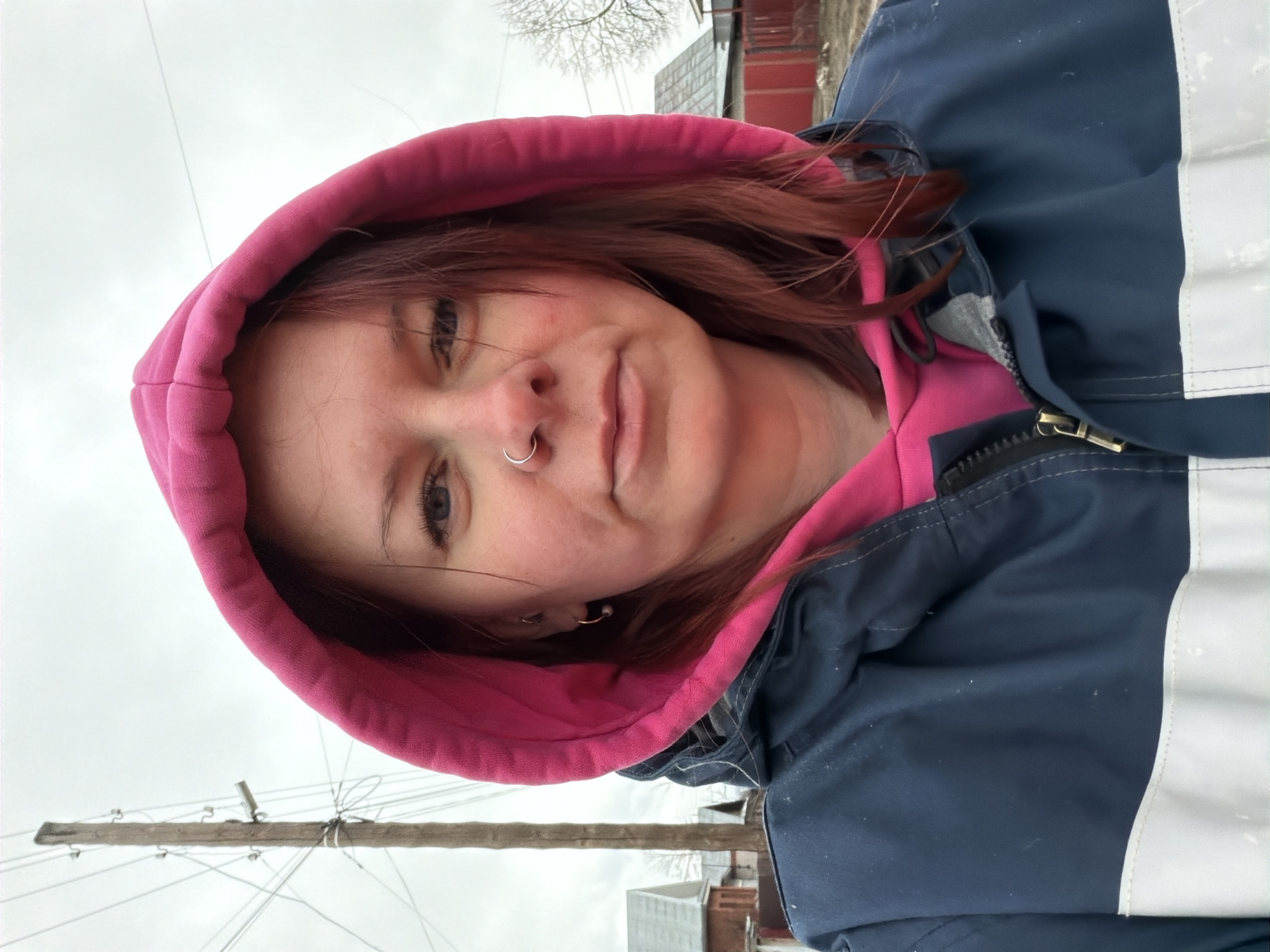 The image shows a woman in a pink hooded sweatshirt underneath a dark blue jacket with white reflective stripes. She has red hair, a nose ring, and an earring. The background suggests an overcast day, with a utility pole and wires visible. Beyond that are hints of buildings, including a red brick structure, suggesting an urban environment. Based on the city provided, the location is Chaikovsky, Russia, but it is not possible to provide more specific details of the location within the city from the image alone.