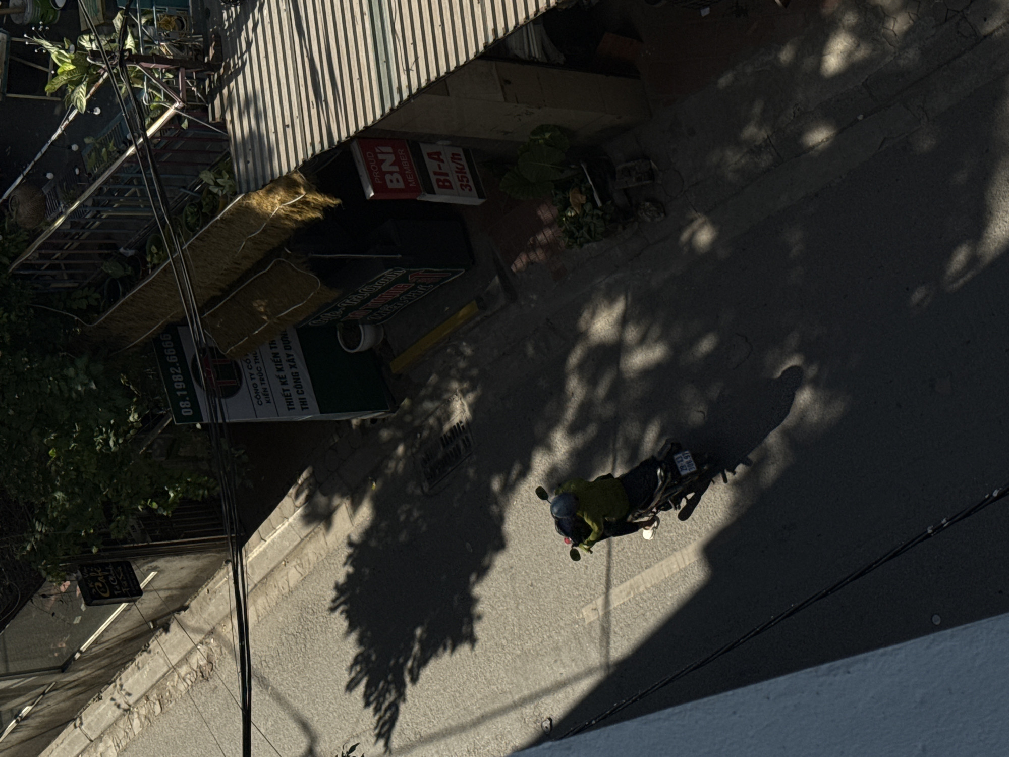 The image is a high-angle shot of a street in what appears to be Hà Nội, Vietnam, captured in daylight. A person on a scooter wearing a helmet is prominently featured on the right side of the frame, navigating the street. The street is shaded by trees, with dappled sunlight filtering through the leaves, creating a pattern of shadows. Buildings line the street, with signs indicating local businesses. A sign that reads "Café Ta" is barely seen hanging on a wall. Another sign advertises a pool hall "BI-A 35k/h", along with "PROUD BNI MEMBER", next to a sign showing "CÔNG TY CỔ KIẾN TRÚC THỦ". Power lines are visible in the foreground.