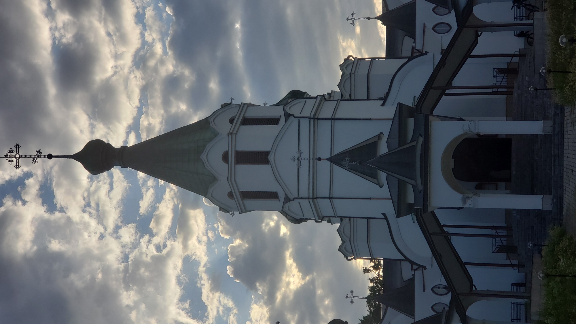 The image depicts the exterior of the Orthodox Cathedral of Saint Nicholas in Prešov, Slovakia, viewed from the front. The cathedral's white facade is partially illuminated by the sun breaking through a dramatic sky filled with clouds, creating a contrast between light and shadow. The central tower, topped with a green dome and a multi-barred cross, dominates the frame. Smaller domes and architectural elements, including arched entrances and covered walkways leading to the main entrance, are visible. Steps ascend to the entrance, and decorative crosses adorn various parts of the building. In the foreground, greenery and paving stones are visible, and faint figures and bicycles can be observed near the entrance area, suggesting a public space. The overall atmosphere is solemn and majestic, enhanced by the dramatic cloud formations.