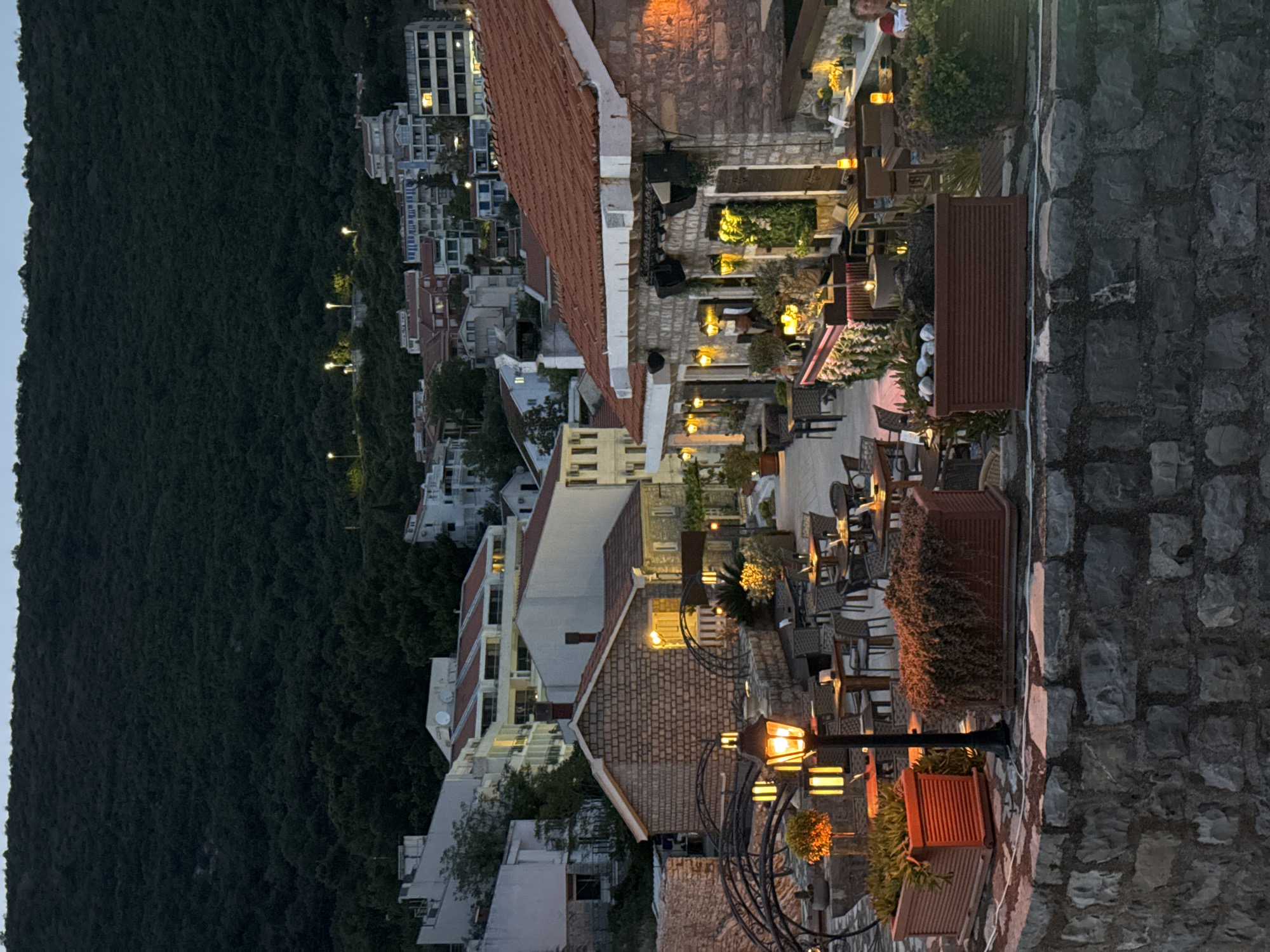 The image shows an elevated, slightly rotated view of Gornji Katun, Montenegro, at dusk. The view is dominated by a steep, forested hillside to the left, with buildings of Gornji Katun situated along the slope and in the valley. The foreground features a stone wall on the right side. Tables and chairs of an outdoor restaurant are spread along the roof tops. The light of lanterns and outdoor lighting creates a warm glow on the rooftops.