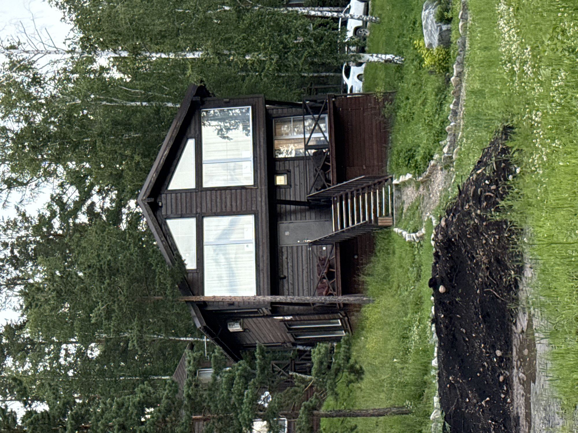 The image shows a two-story wooden house situated on a grassy slope surrounded by a dense forest. The house has a dark brown exterior and large windows. A set of wooden stairs leads up to a balcony on the second floor. In the foreground, there is a pile of dark soil and a small puddle. White cars are visible parked in the background. The scene appears to be a rural or semi-rural setting, potentially a vacation home or a residential property in Syrostan, Russia, given the architectural style and surrounding vegetation, which is consistent with the region's natural environment. No people are visible in the image.